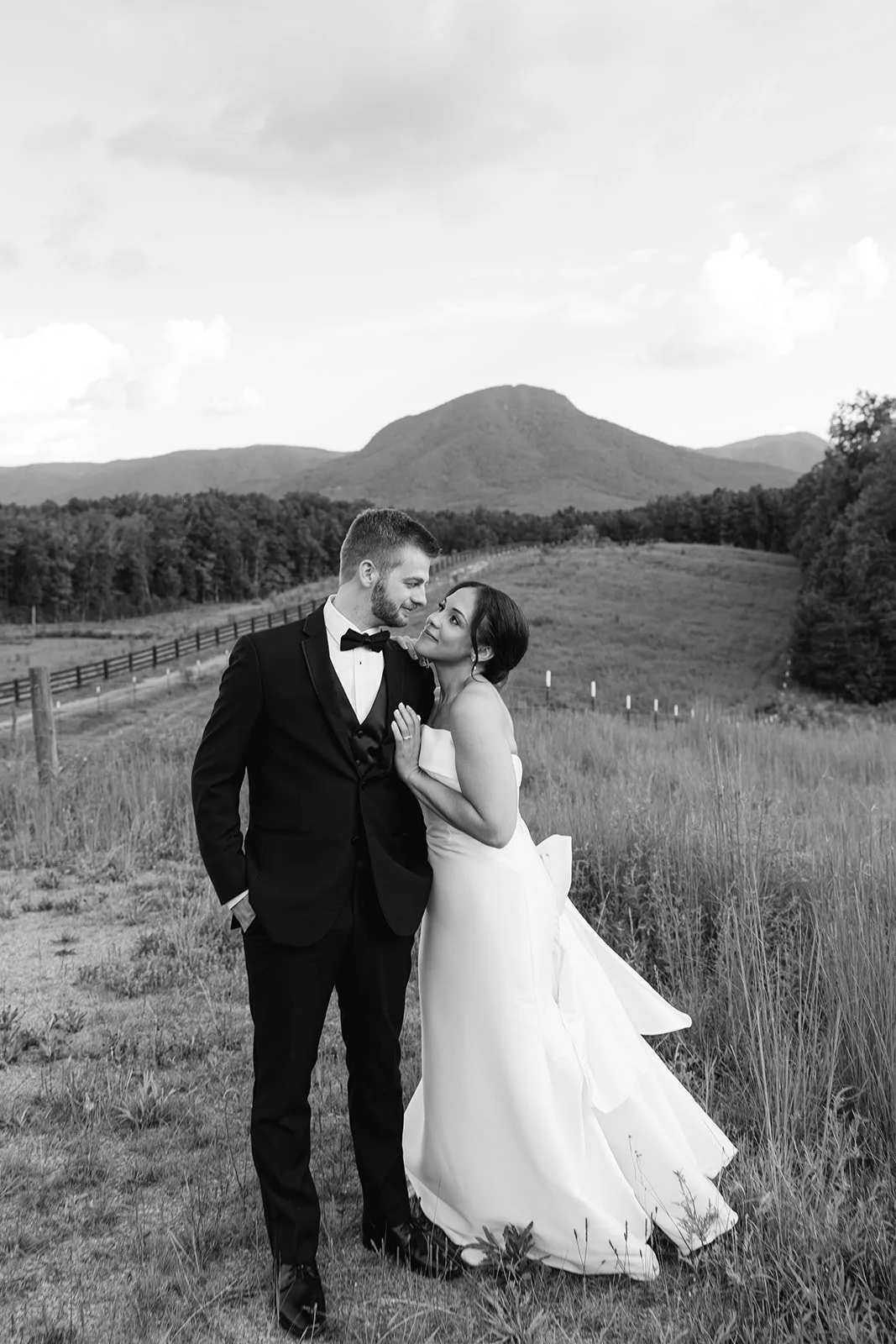 A black and white photo of a bride and groom standing close in a grassy field, with mountains and a partly cloudy sky in the background. The groom is wearing a tuxedo, and the bride is in a strapless wedding gown, looking lovingly at each other.