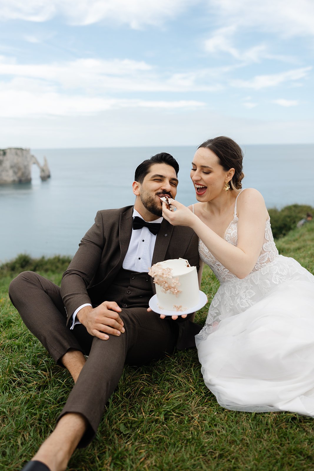 Bride and groom in wedding attire sitting on grass, sharing cake outdoors near a scenic body of water and cliffs.