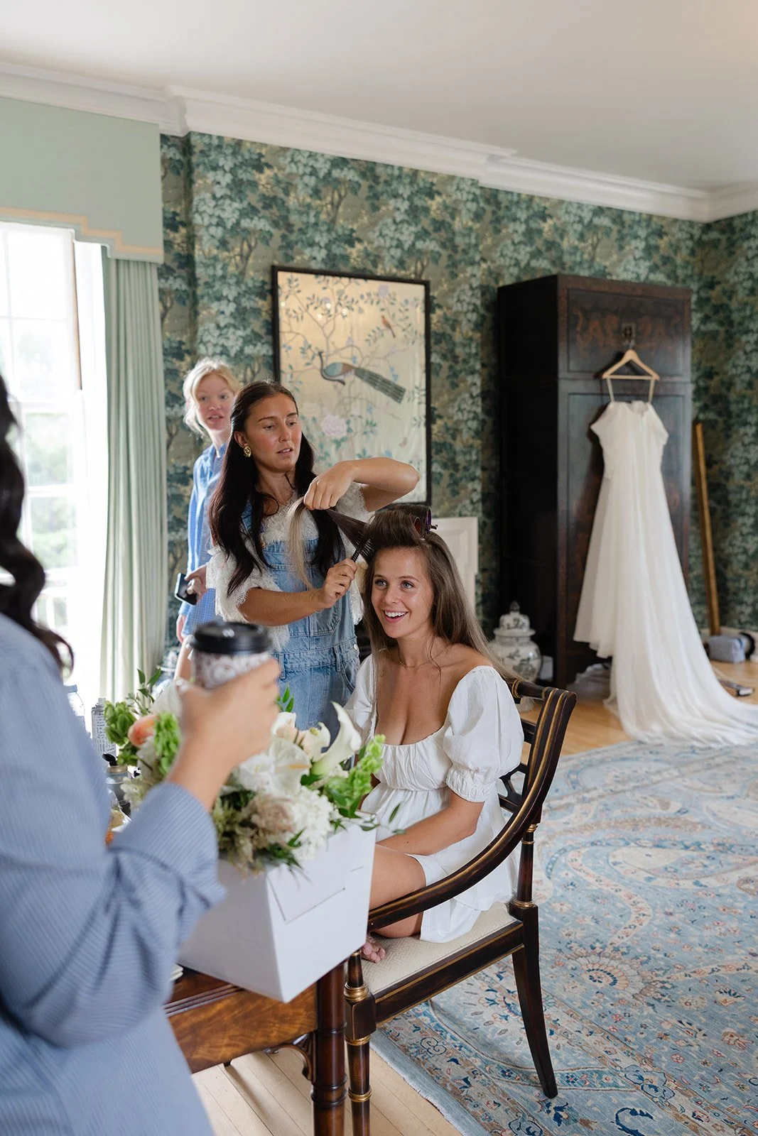 A woman getting her hair styled by a hairdresser while another woman holds a coffee cup, with a wedding dress hanging in the background.