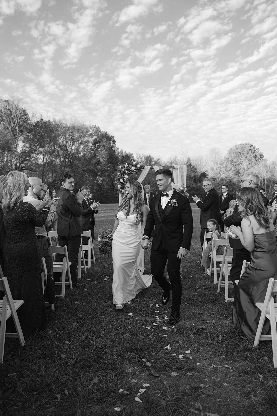 A black-and-white wedding ceremony outdoors with a bride and groom walking hand in hand down the aisle, surrounded by seated and clapping guests, under a partly cloudy sky.