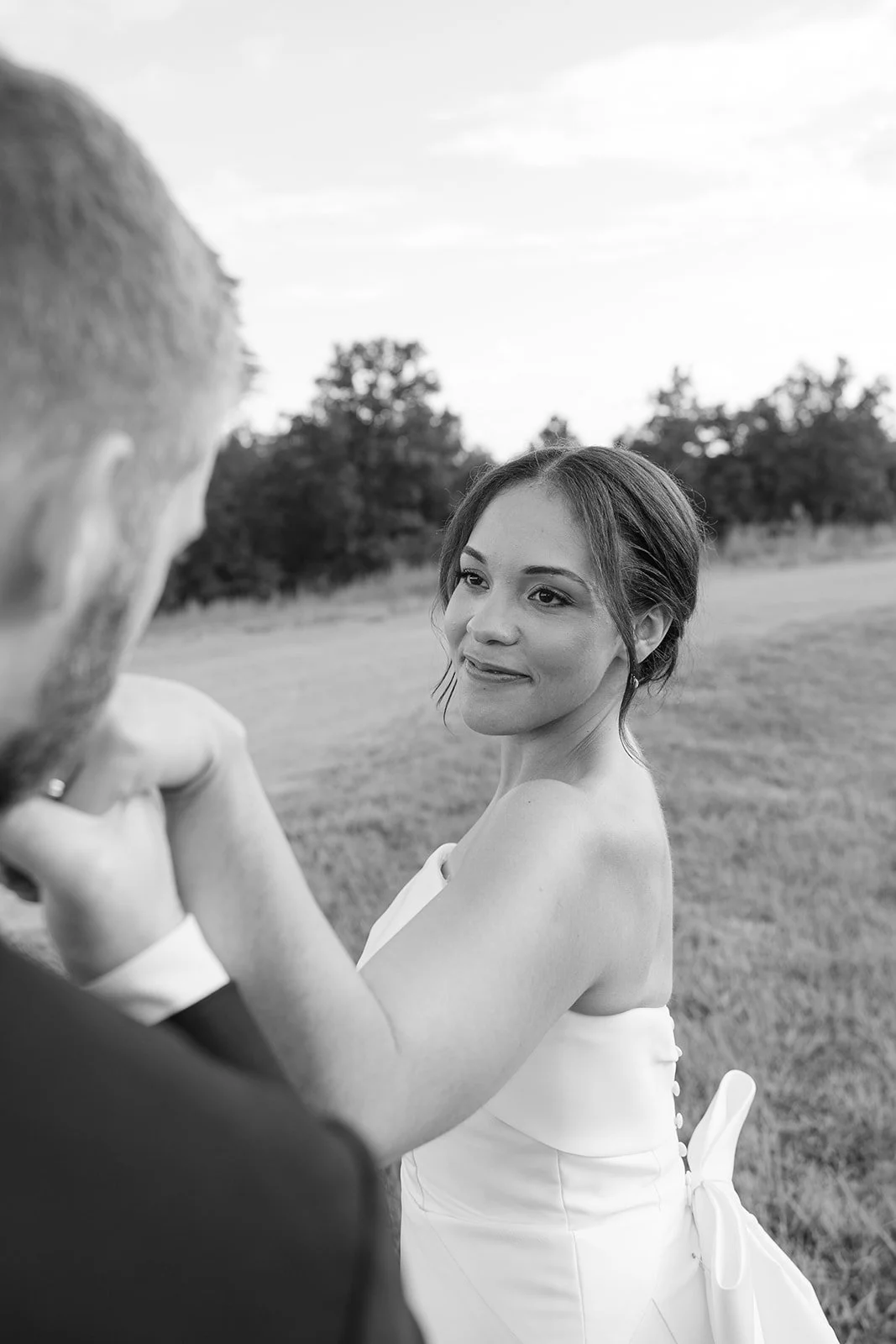 A woman in a strapless white dress looks at a man, outdoors in a field with trees in the background, during sunset or early evening.
