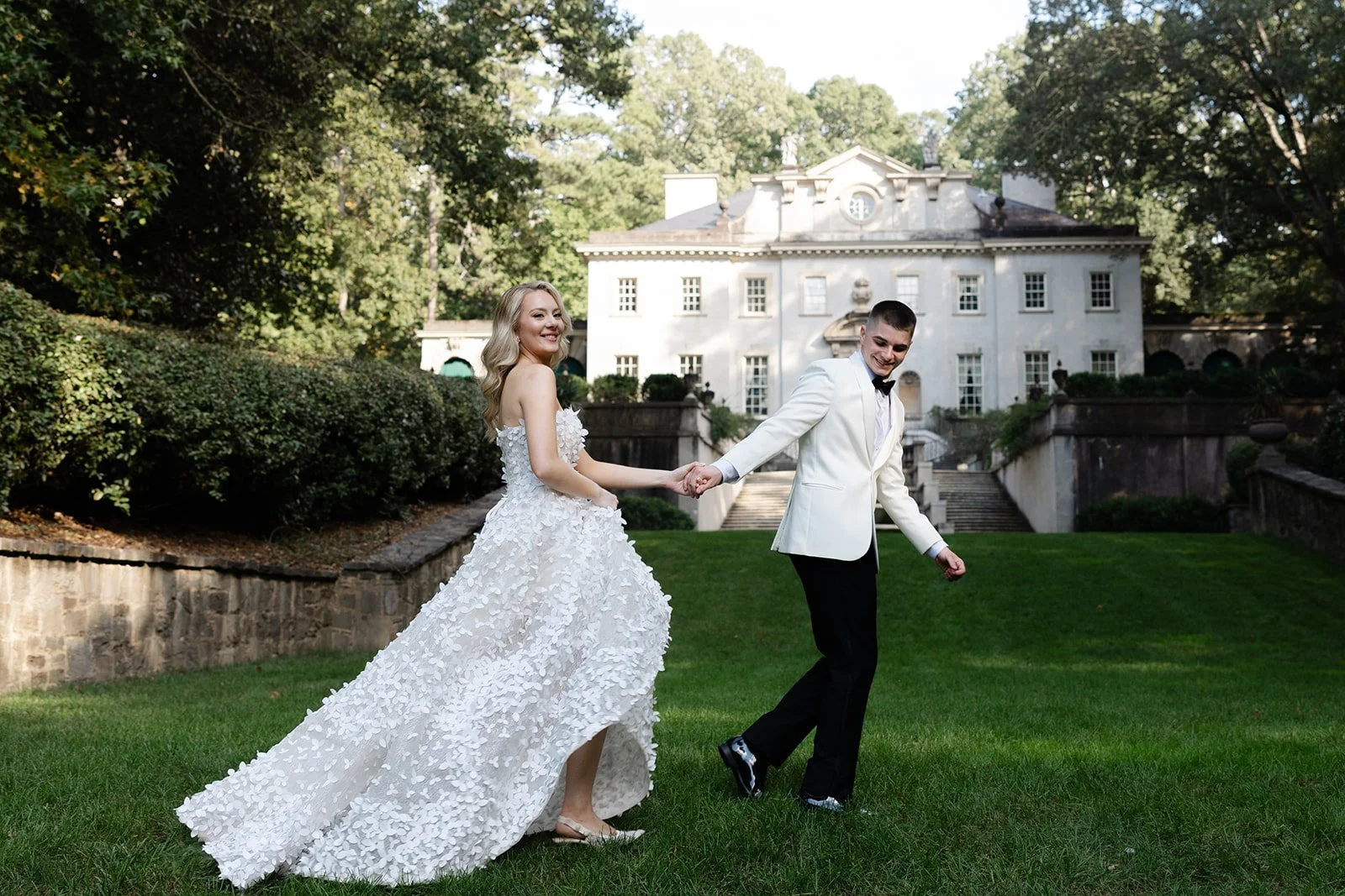 A bride and groom holding hands and walking on a lawn outside a large white mansion surrounded by trees, with the bride in a white wedding dress and the groom in a tuxedo.