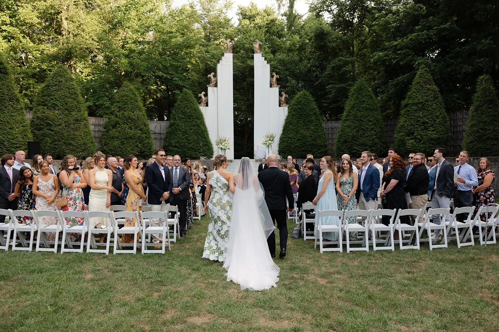 A wedding ceremony taking place outdoors on a grassy area, with a bride walking down the aisle accompanied by a person, possibly her father. Guests are seated on white chairs on either side of the aisle, watching the bride approach. The setting inclu