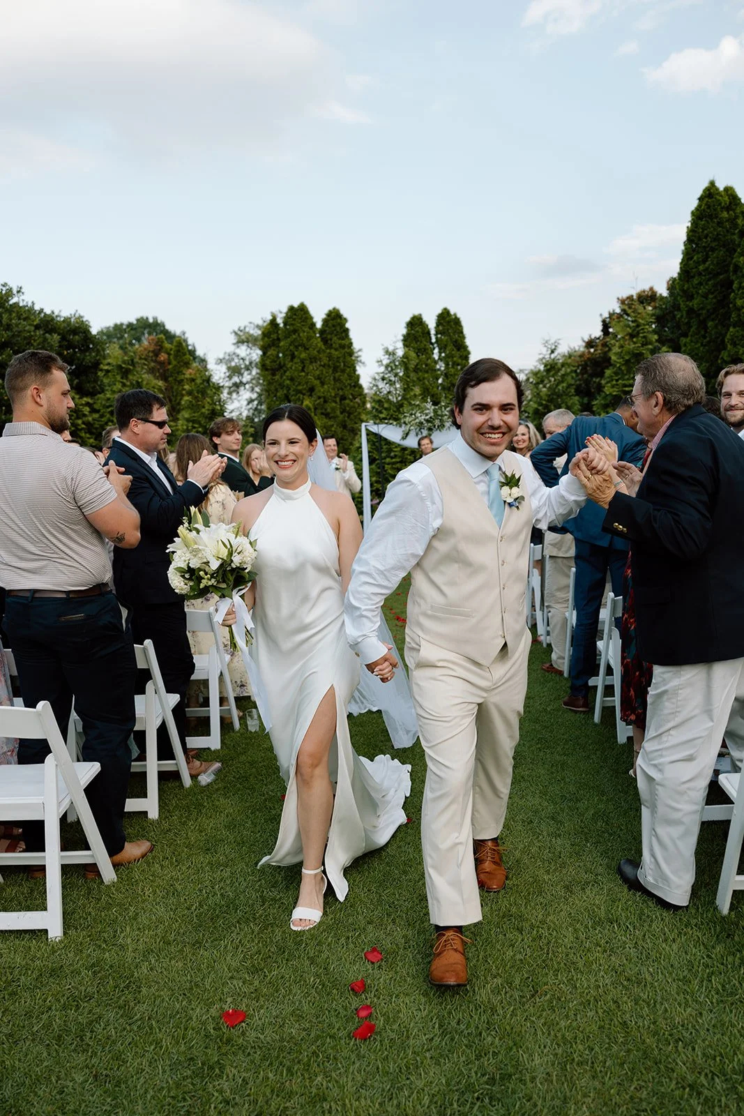 A newlywed couple walking hand in hand down the aisle at their outdoor wedding, smiling. The bride is holding a bouquet of white flowers, wearing a white dress with a thigh-high slit. The groom is dressed in a light-colored vest and pants, with a lig