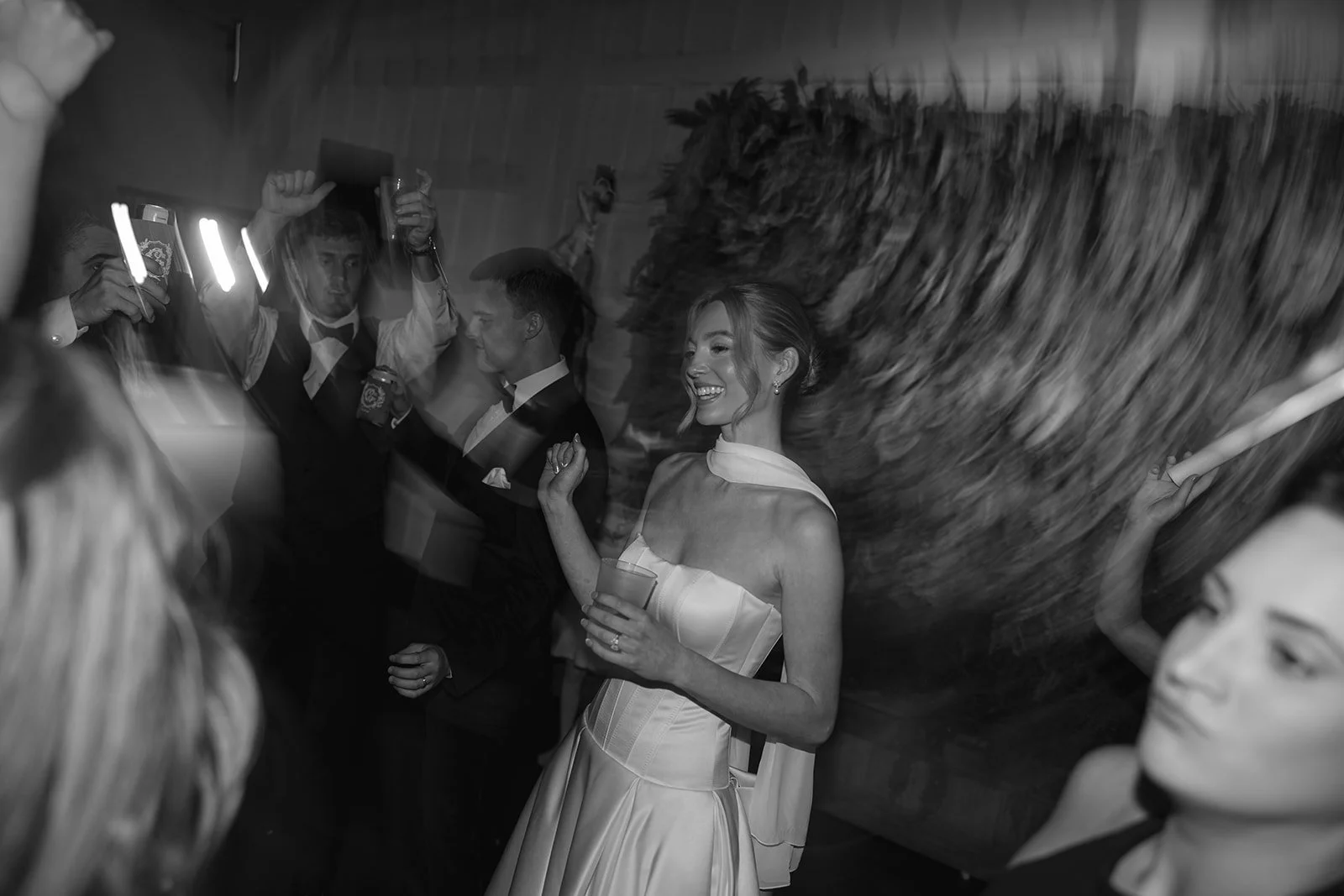 People dancing and celebrating at a party or wedding reception, with a woman smiling holding a drink in a strapless dress, black and white photo.