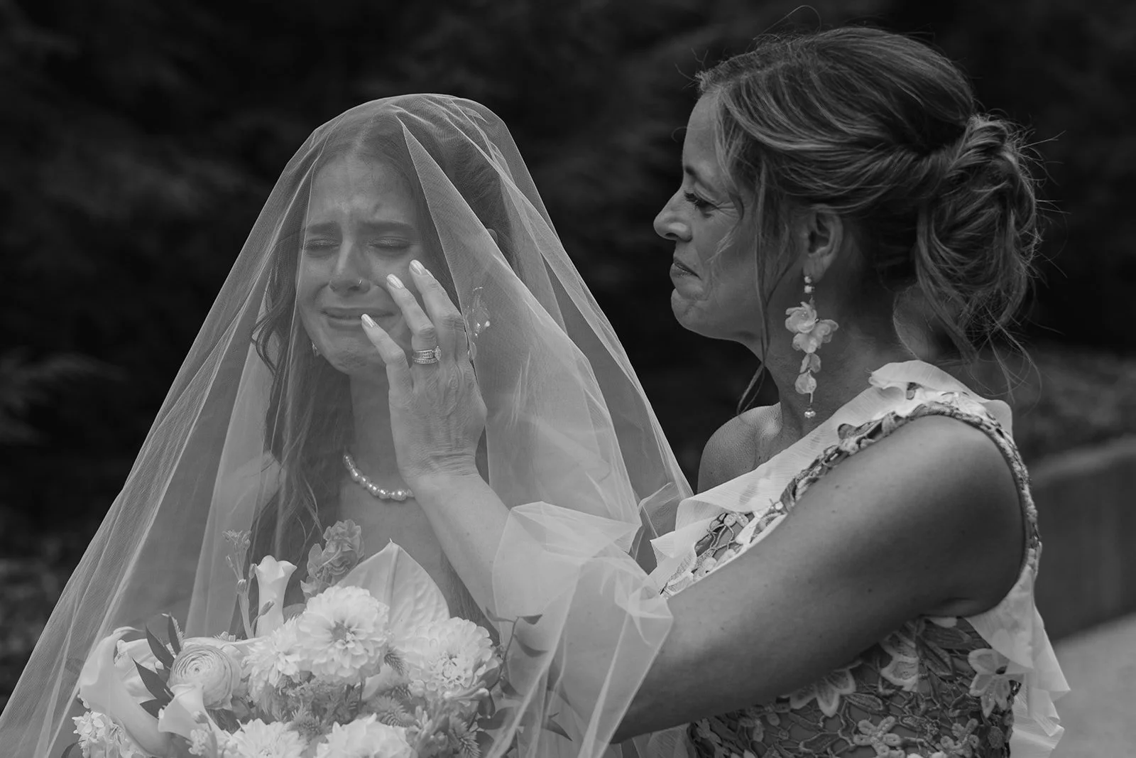 A woman dressed in a formal attire with flowers and earrings is shown comforting a crying bride wearing a veil and holding a bouquet of flowers.