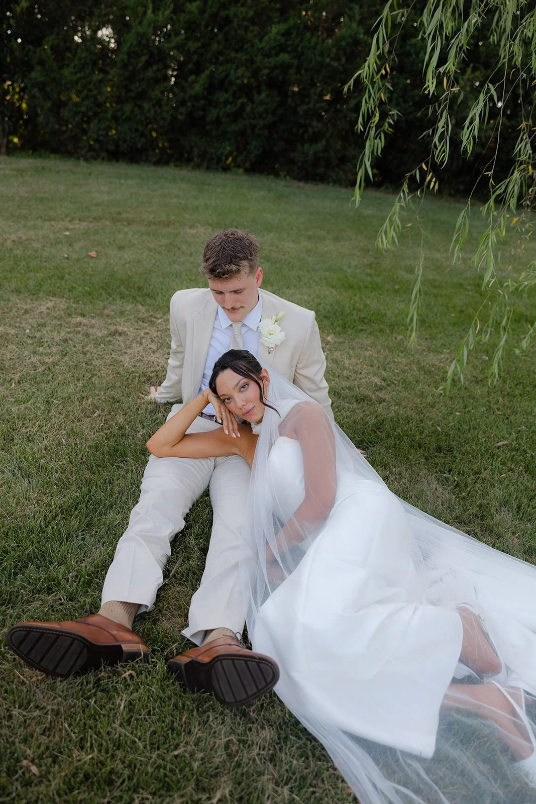 A bride and groom sit on the grass after their wedding, with the bride leaning on the groom's lap and the groom looking down at her, in a park-like setting with trees and greenery in the background.