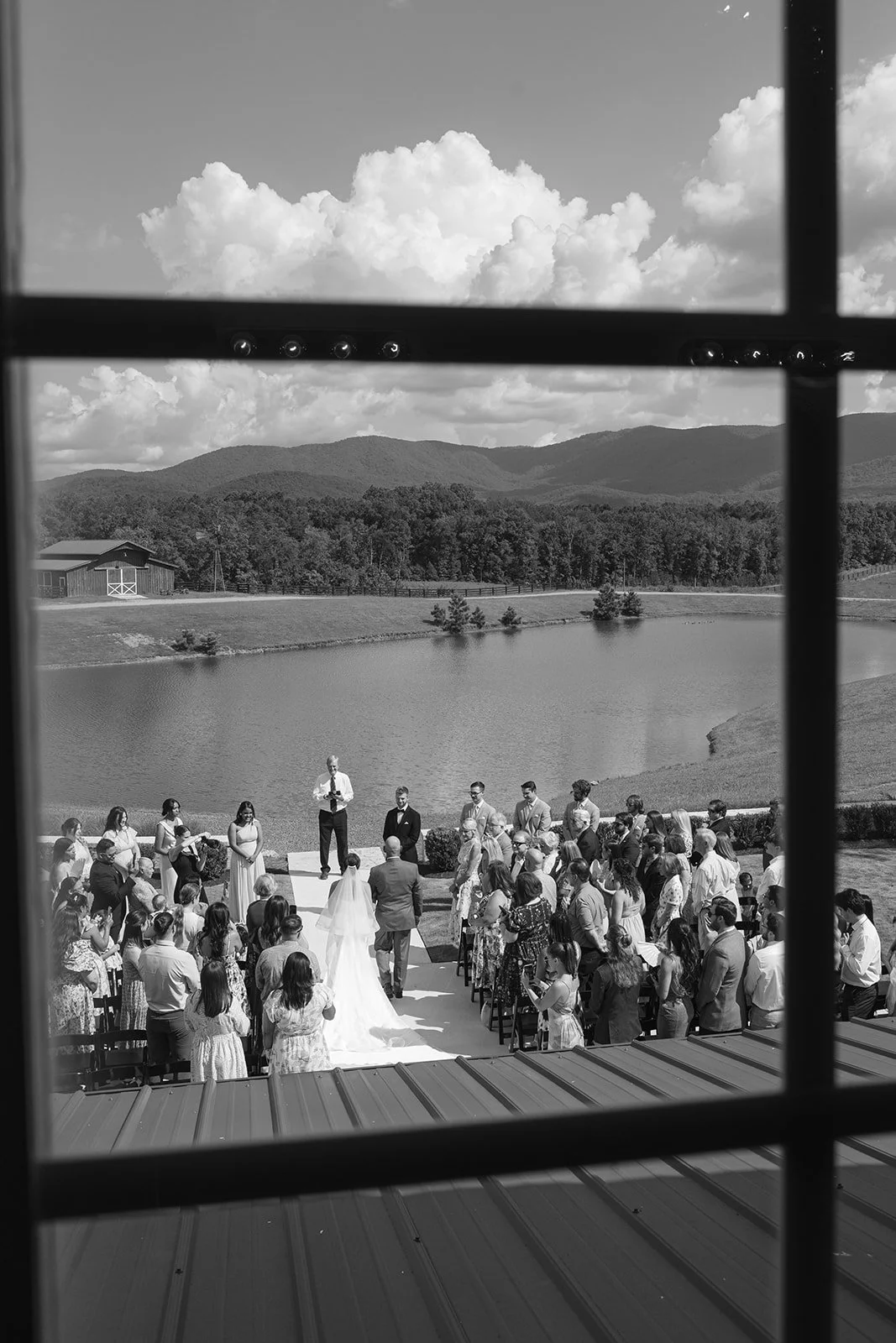 Black and white photo of a wedding ceremony outdoors near a lake. View seen through a window with grid panes. The bride and groom are standing at the altar with officiant, surrounded by guests.