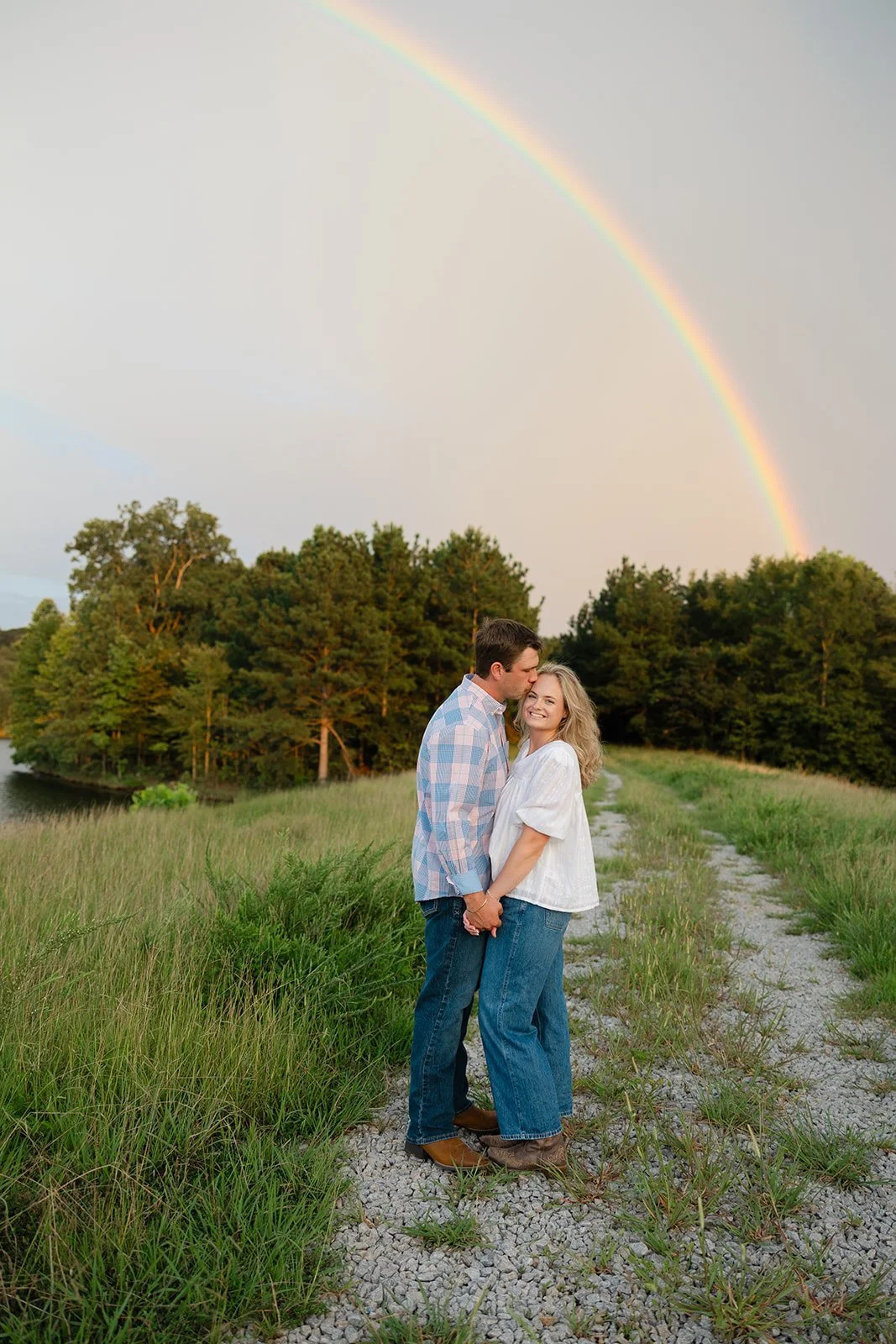 A couple standing on a gravel path, holding hands, smiling, with a rainbow in the sky above them, green trees in the background, during sunset.