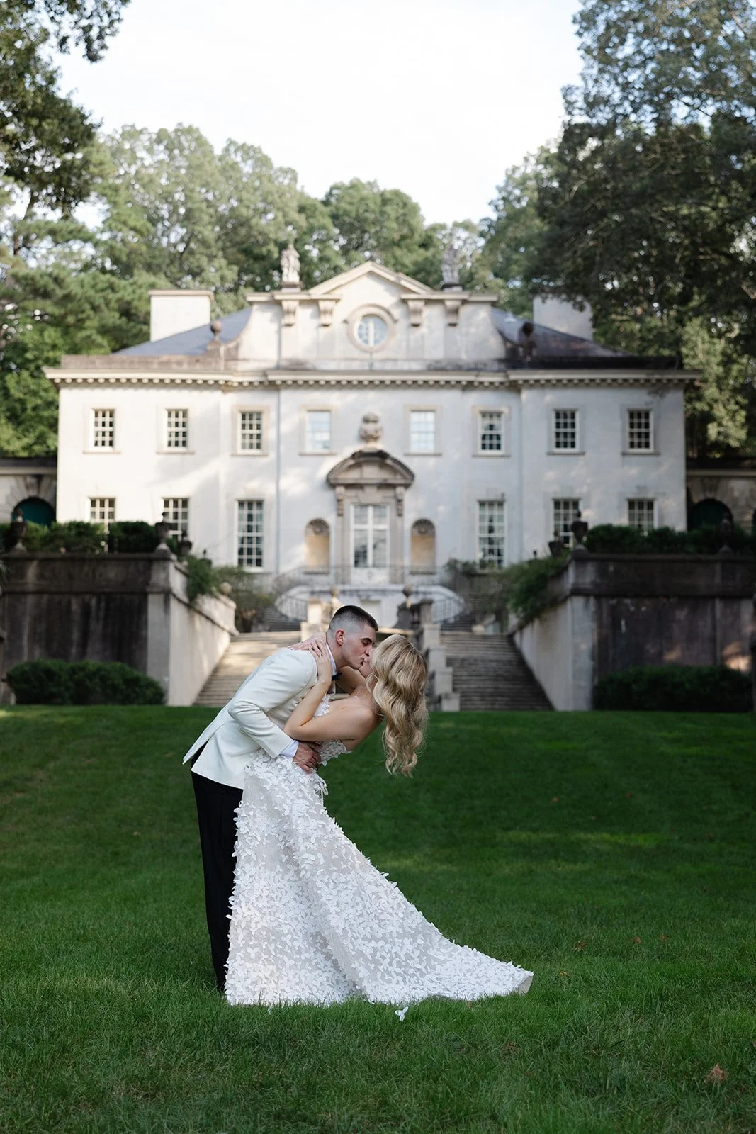 A bride and groom share a kiss on a lush green lawn in front of a grand, historic mansion.