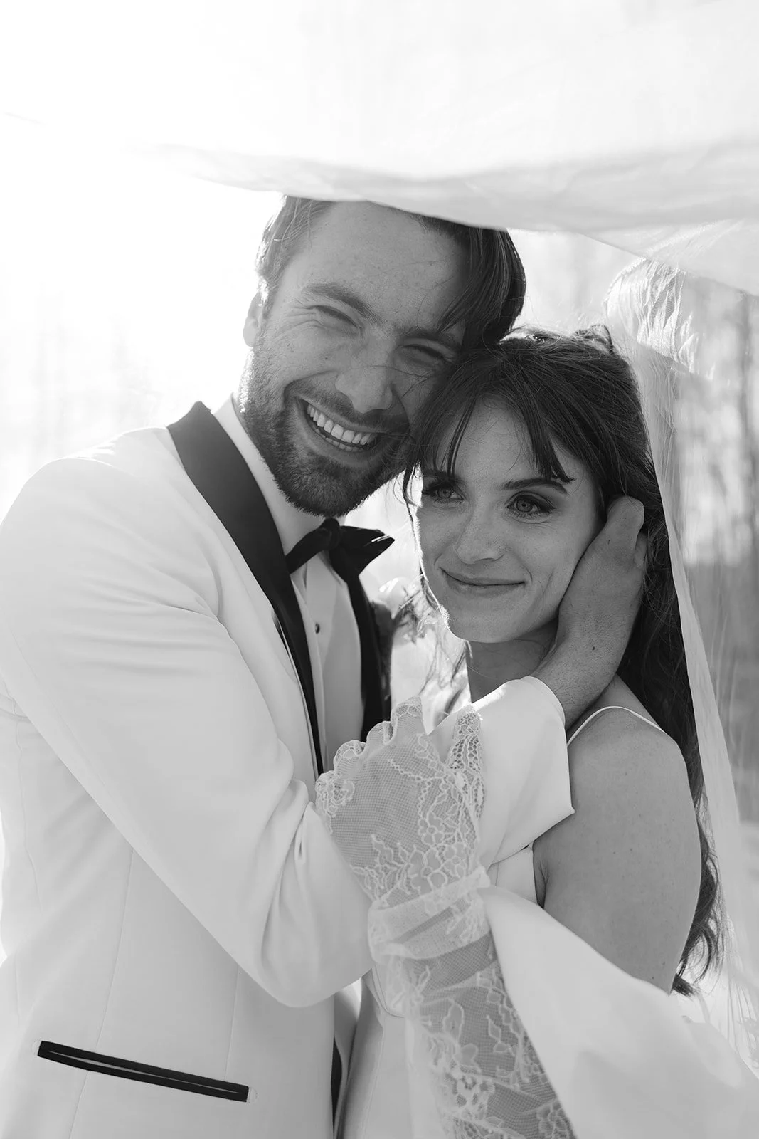 Black and white photo of a happy groom and bride embracing, with the groom smiling widely and the bride gently smiling, under a veil or fabric canopy.