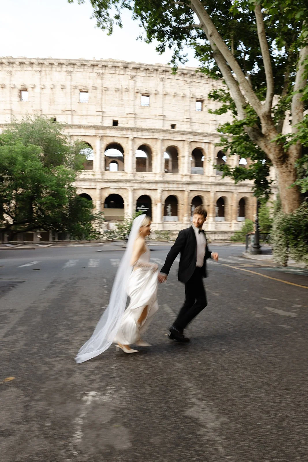 Bride and groom holding hands, crossing a street in front of the Colosseum in Rome, Italy, with trees on both sides.