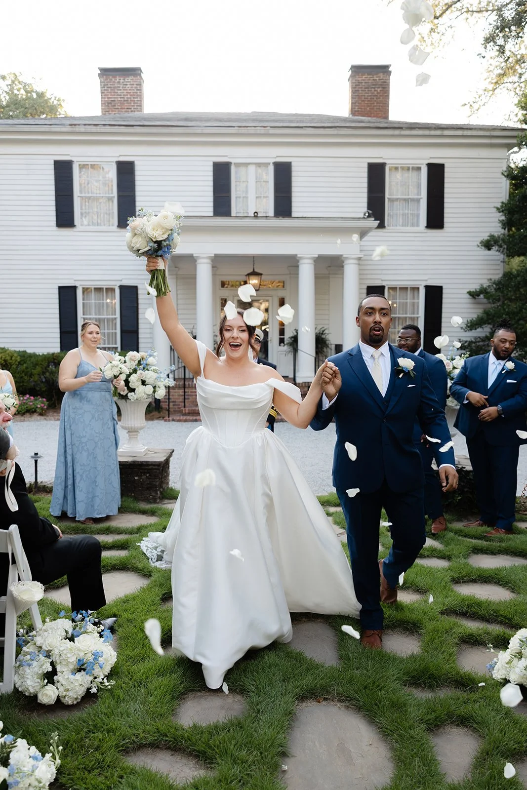 A bride and groom celebrating during their wedding ceremony outside a white house. The bride, in a white gown, is smiling and holding a bouquet of flowers high in the air as flower petals fall around them. The groom is in a dark blue suit, holding the bride's hand, with other wedding guests in the background.
