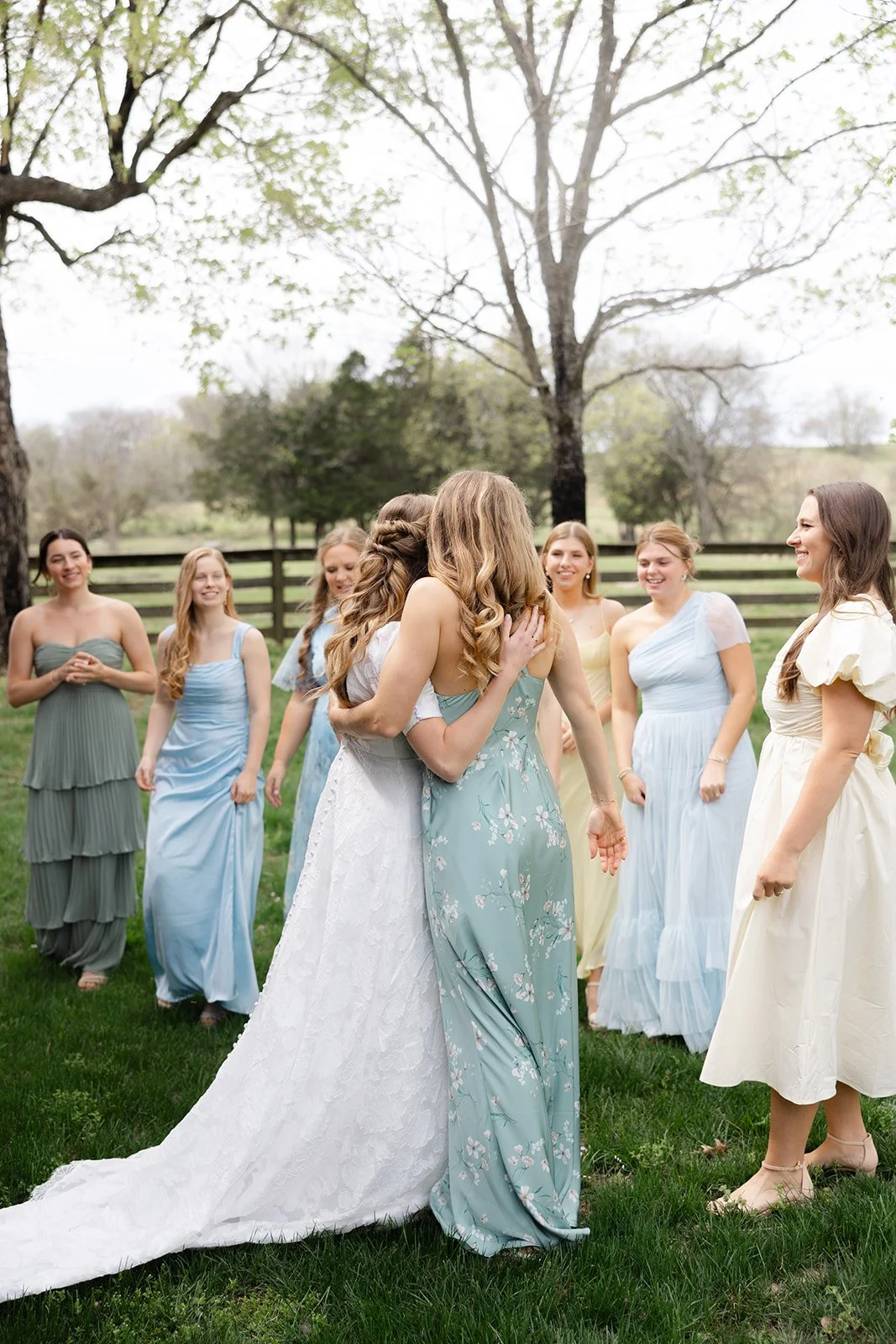 A bride in a white wedding dress hugs a woman in a blue floral dress among a group of women outside in a grassy area with trees and a wooden fence.