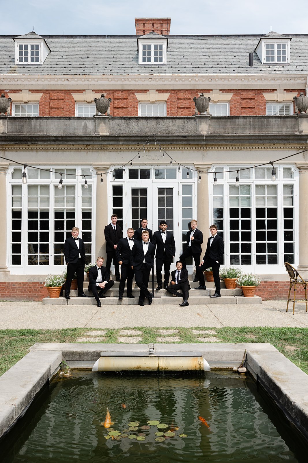 A group of nine men in tuxedos posing in front of a historic building with large windows, plants in pots, and string lights, outside near a fountain with koi fish.