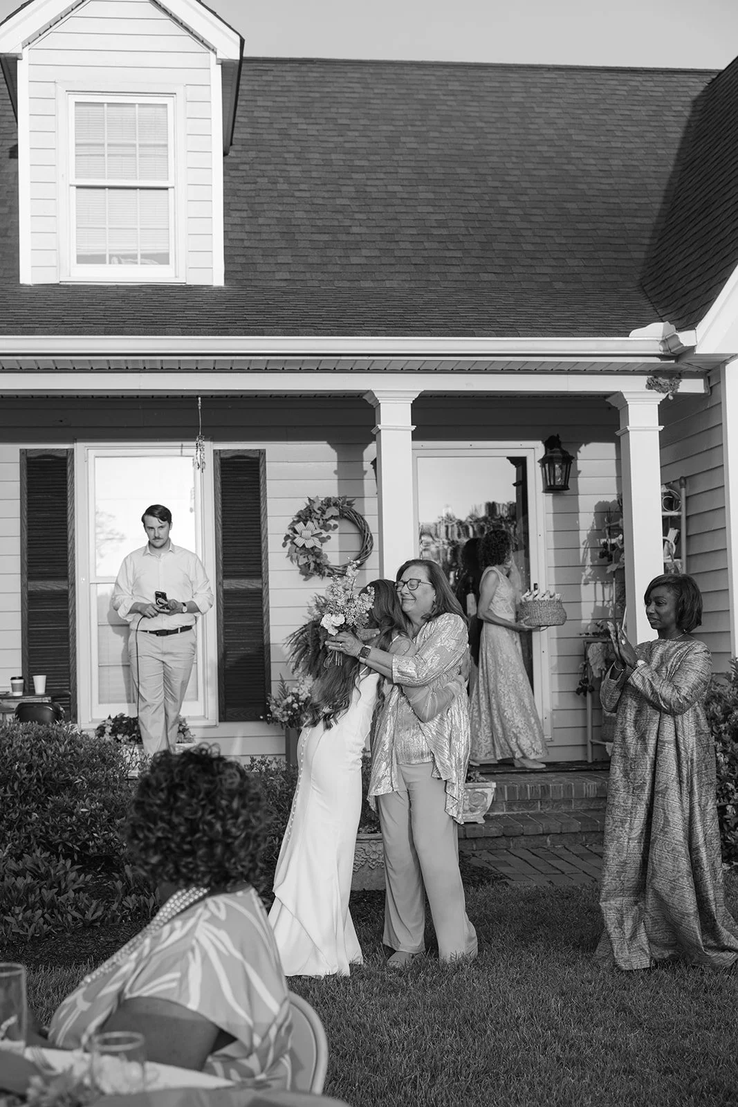 A group of people—women, a young girl, and a man—gather outside a house for a celebration. An older woman and a young girl embrace in the foreground, while others are engaging in conversation or taking photos. The house has a porch with decorations l