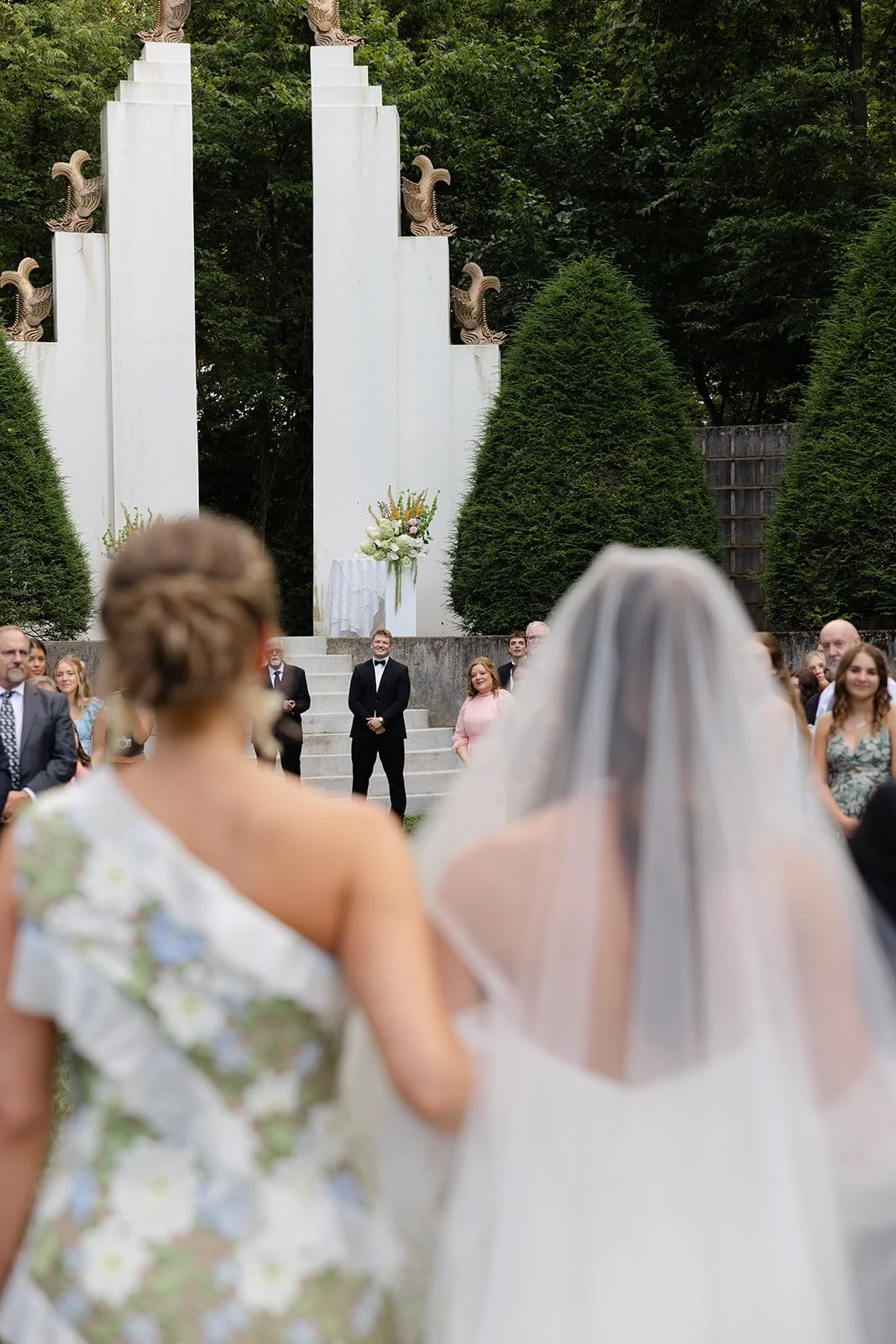 A wedding ceremony outdoors with guests standing and a bride in a white veil and dress facing a white altar with floral arrangement, surrounded by tall trees.