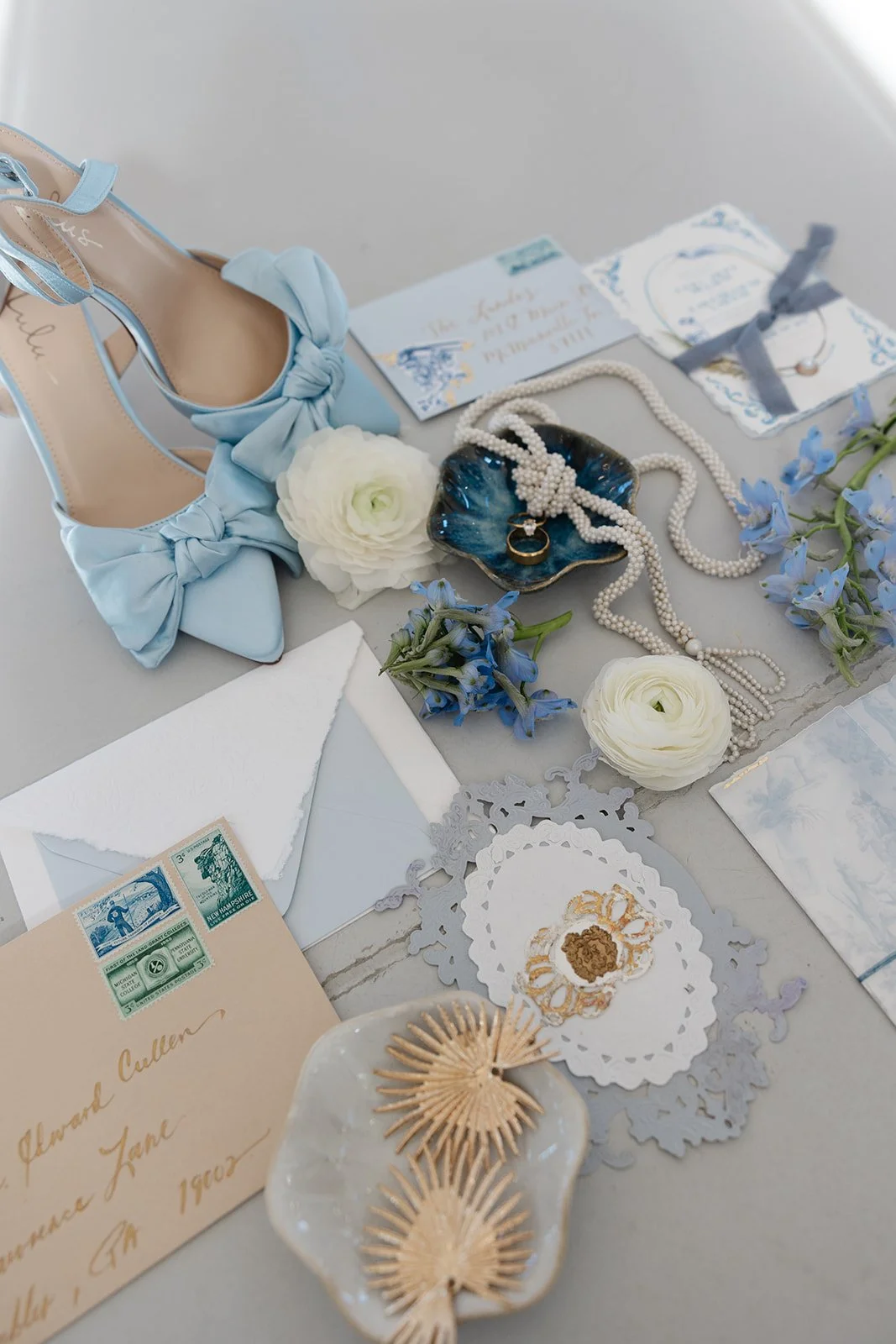 A flat lay of vintage wedding items including blue high heels with bows, white flowers, pearl necklaces, rings, handwritten notes, vintage stamps, and ornate lace paper.