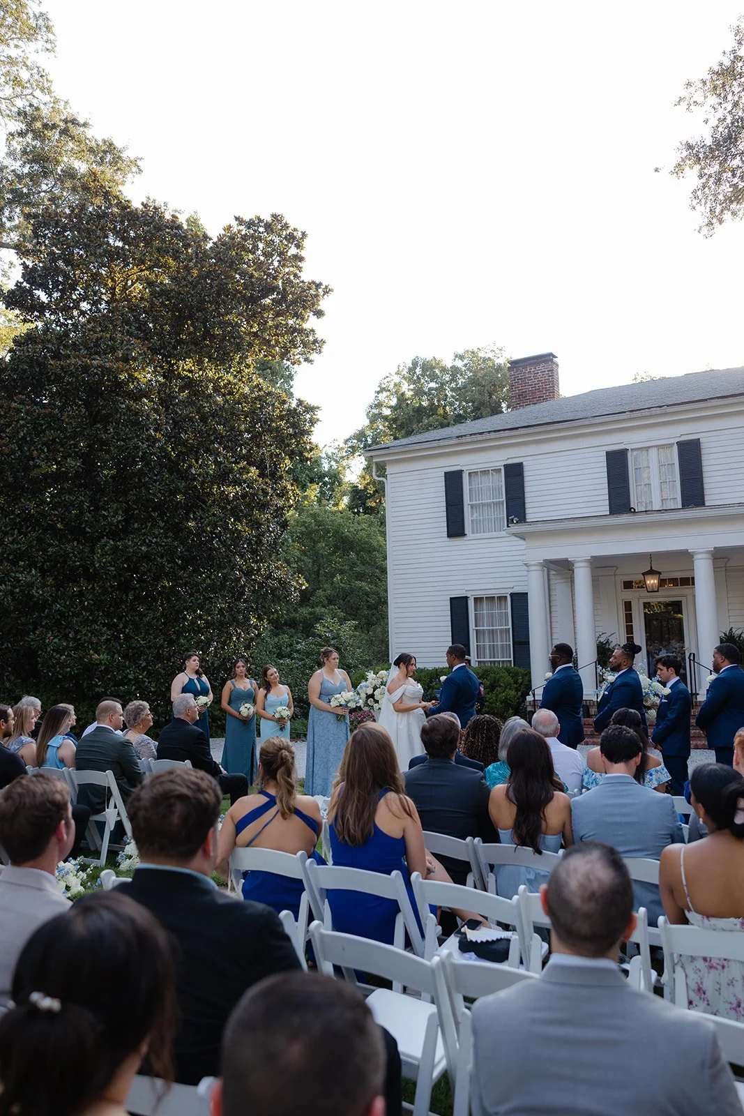 Wedding ceremony taking place outdoors in front of a white house with black shutters. The bride and groom are exchanging vows, surrounded by bridesmaids and groomsmen. Guests seated in white chairs watch the ceremony.