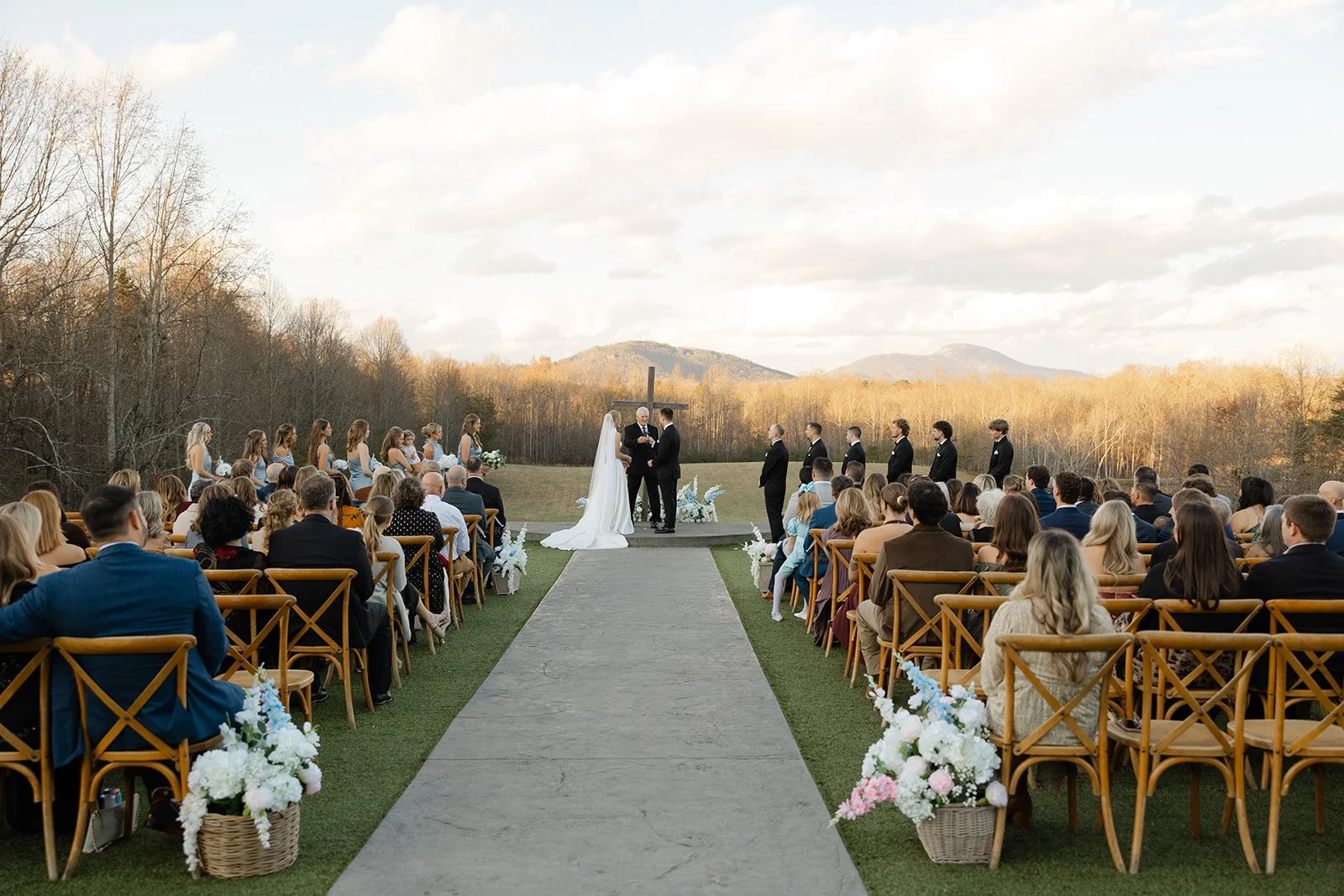 Outdoor wedding ceremony with a bride and groom at the altar, surrounded by bridal party and guests, in a scenic natural setting with mountains in the background.