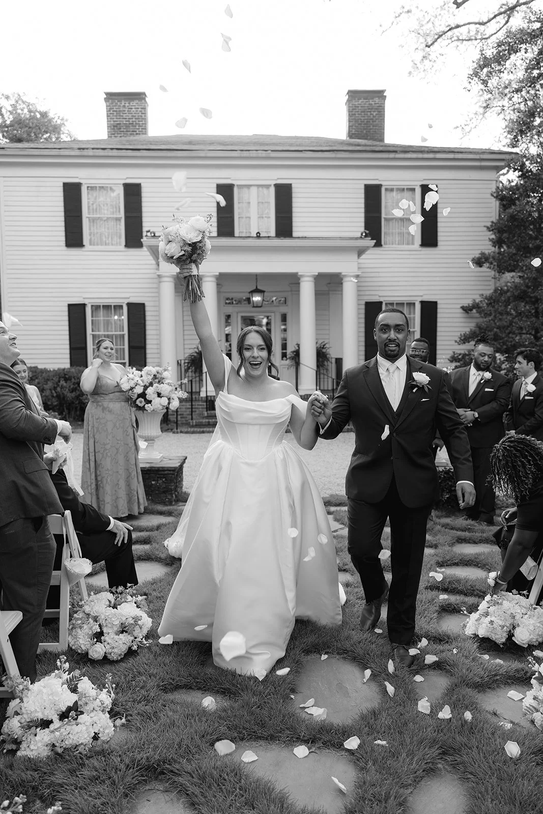 A newly married couple walks hand-in-hand celebrating outside a house, with rose petals falling around them. The bride is holding a bouquet and smiling, while the groom is dressed in a suit. Guests are gathered around them, and some are throwing flow