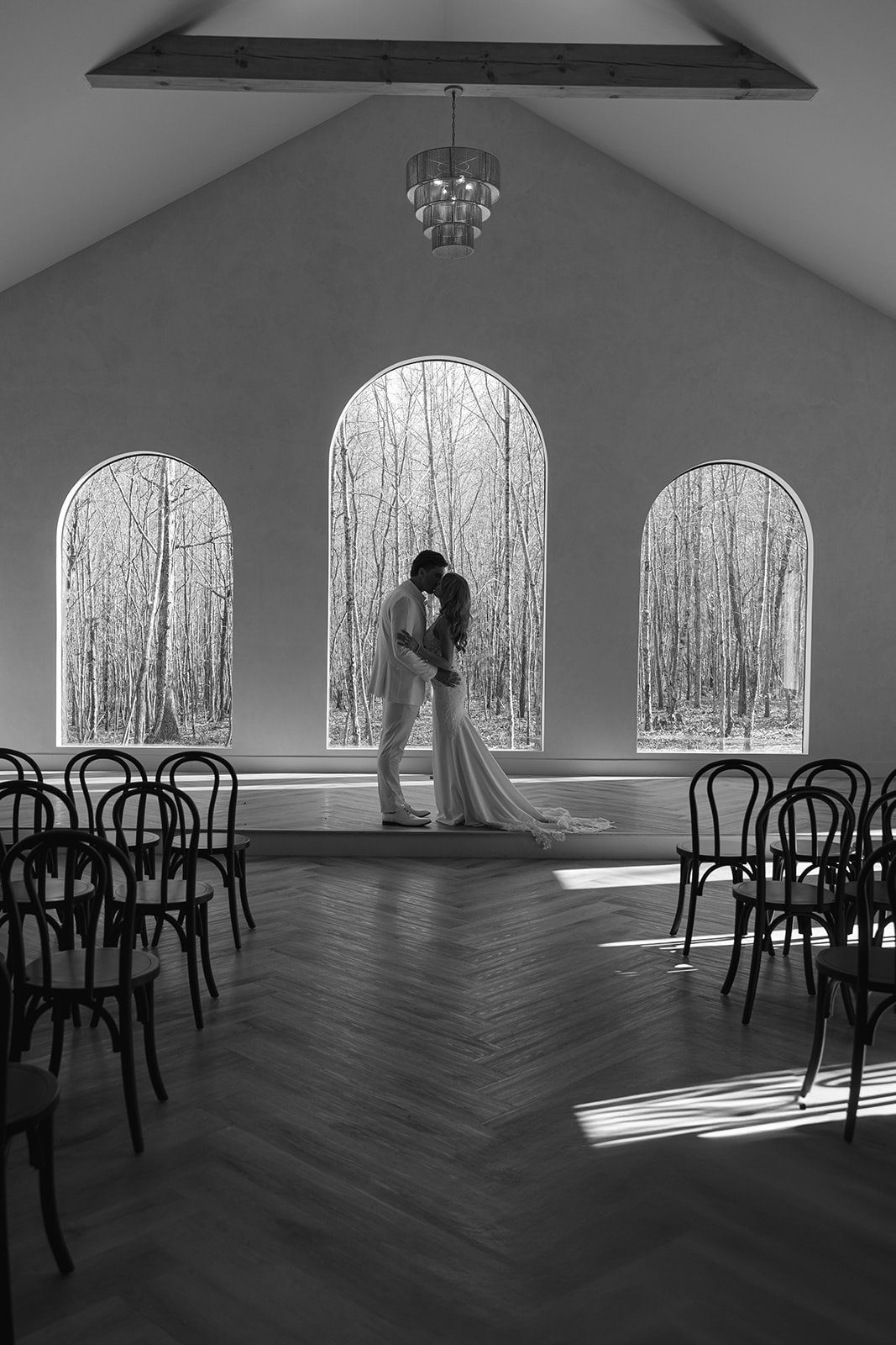 A couple in wedding attire sharing a kiss inside a chapel with large arched windows showing a forest outside.