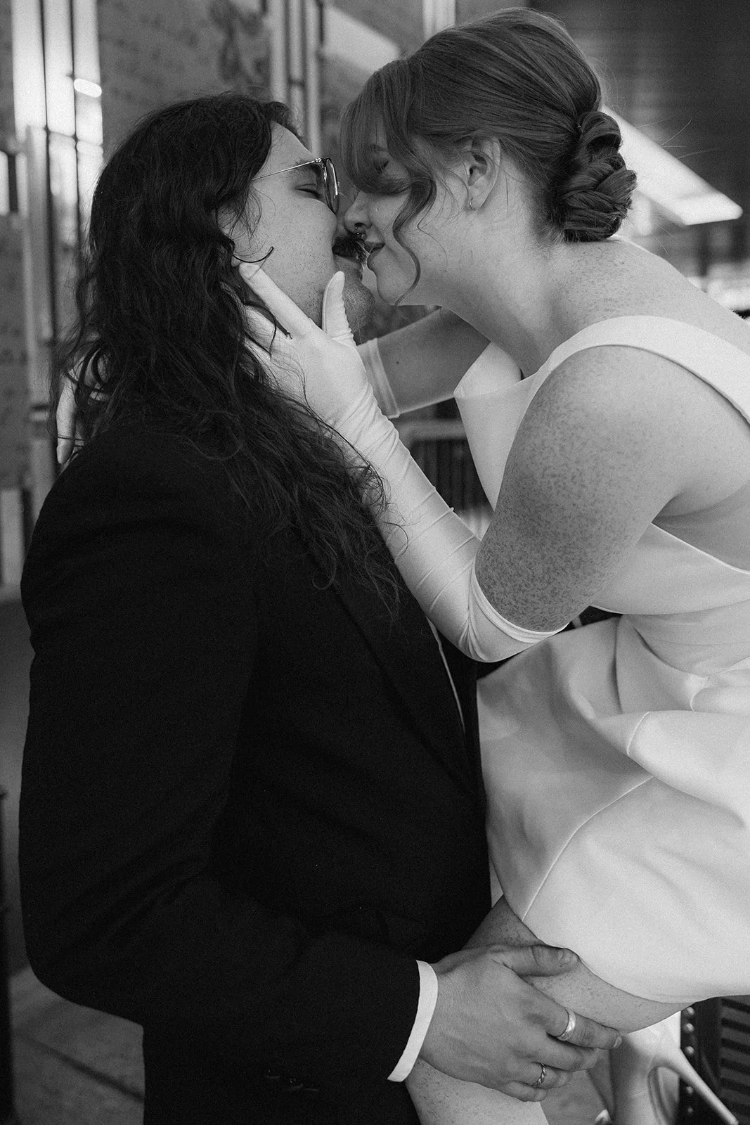 A couple dressed in formal wedding attire sharing an intimate moment, about to kiss, with the bride holding the groom's face and the groom supporting the bride's leg.