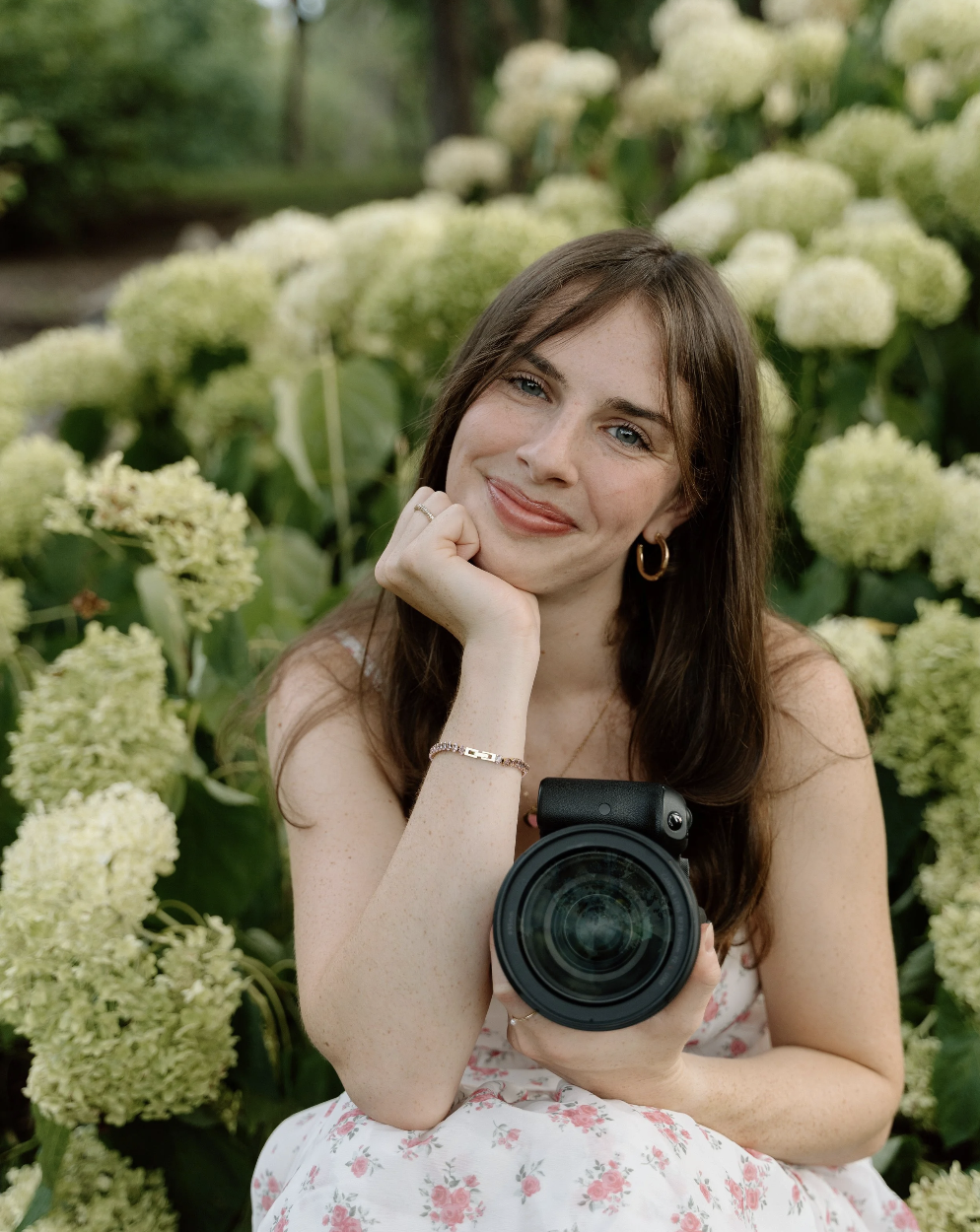A young woman with brown hair and blue eyes, smiling while holding a camera, sitting among white hydrangea flowers.