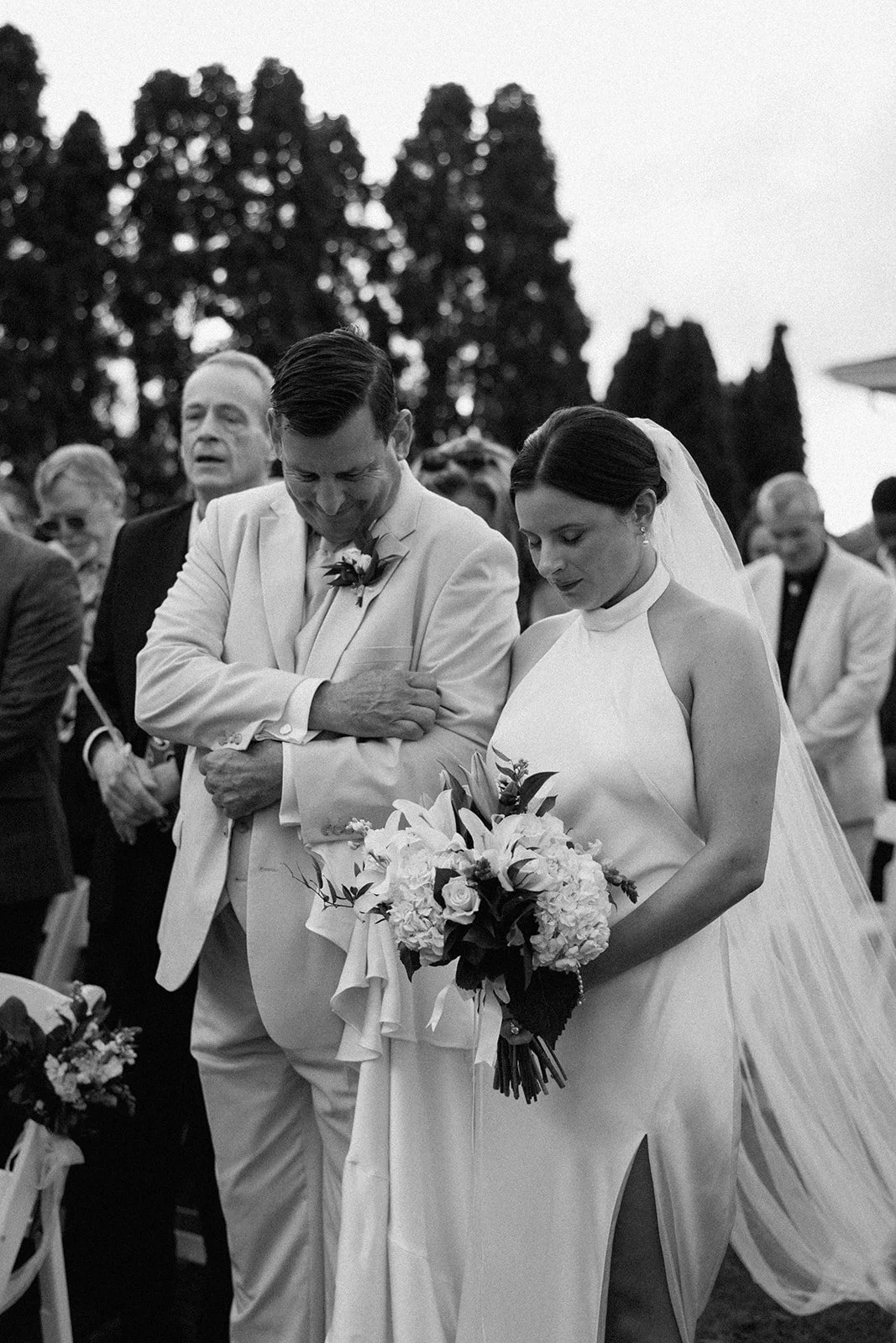 A black and white photo of a bride and groom standing together during a wedding ceremony, with the bride holding a bouquet of flowers and a veil, and the groom wearing a light-colored suit, surrounded by guests in formal attire.