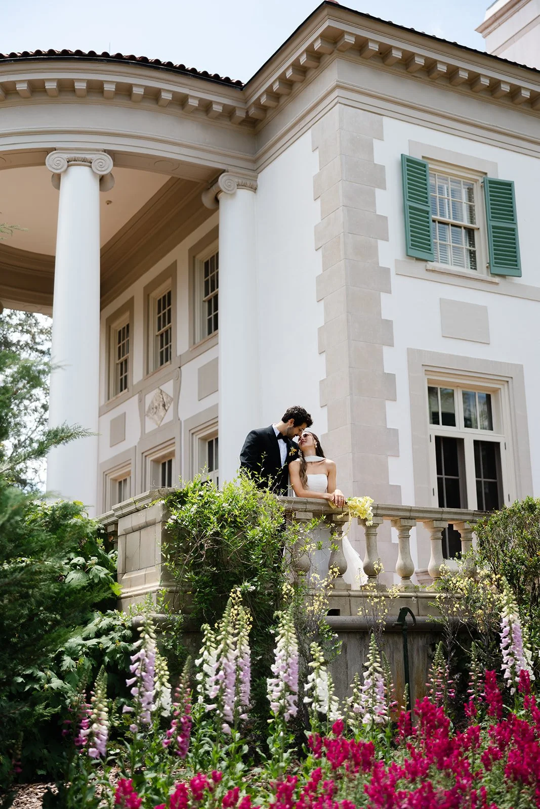 A bride and groom standing on a balcony of a large, elegant white mansion, surrounded by colorful flowers and greenery.