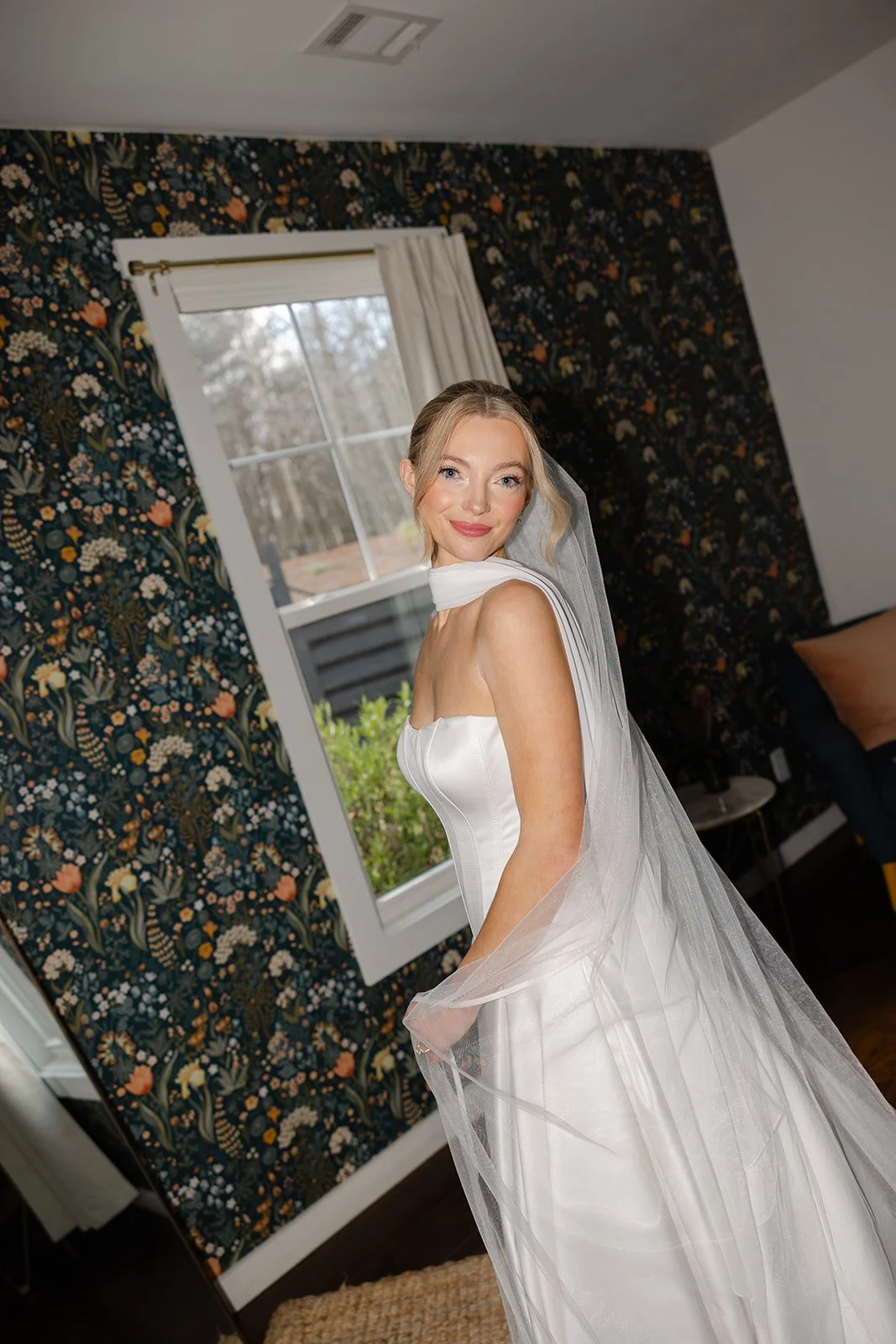 A bride in a white wedding gown with a veil standing indoors near a window, smiling at the camera.