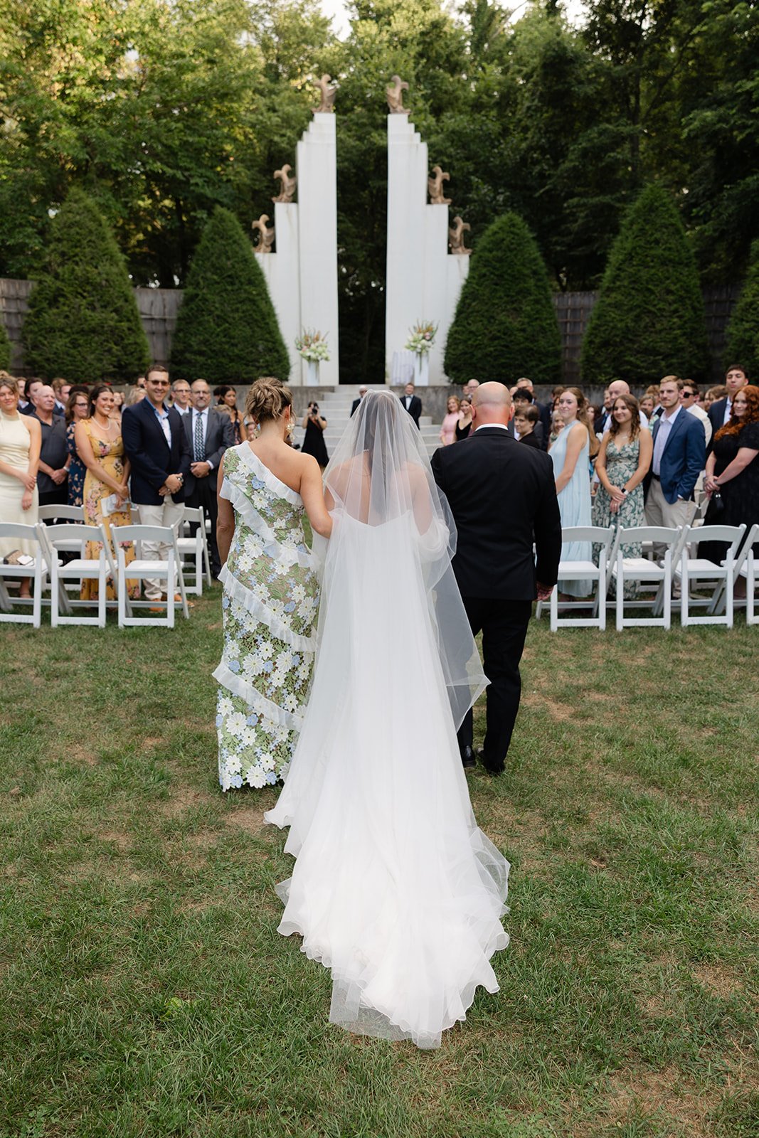 Bride walking down the aisle with her parents at an outdoor wedding ceremony, with guests seated on either side and decorative sculptures and greenery in the background.