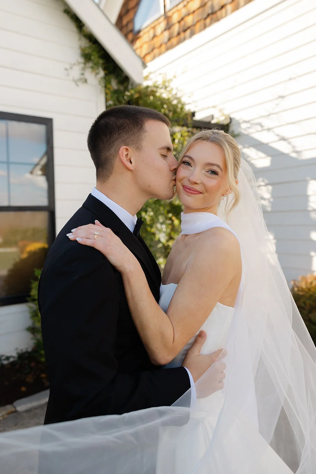 A newlywed couple with the groom kissing the bride on the cheek, smiling outdoors near a white house with green plants.