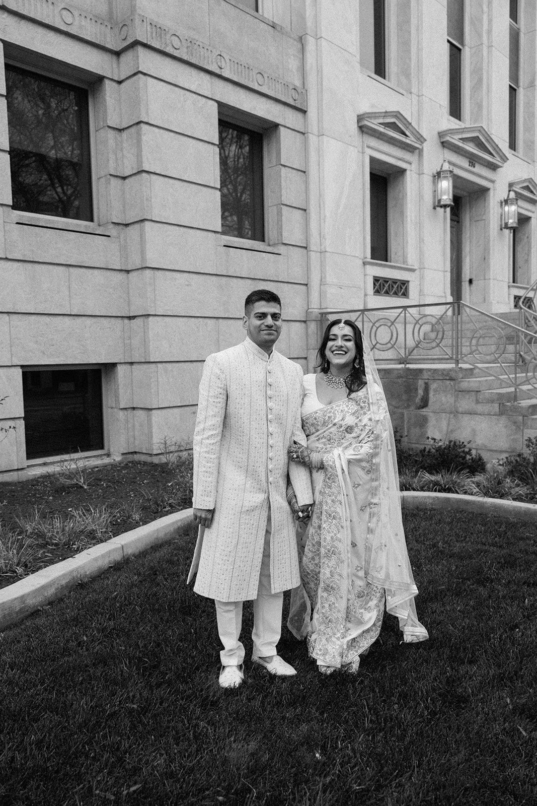 A couple in traditional Indian wedding attire standing on grass outside a building, holding hands and smiling.