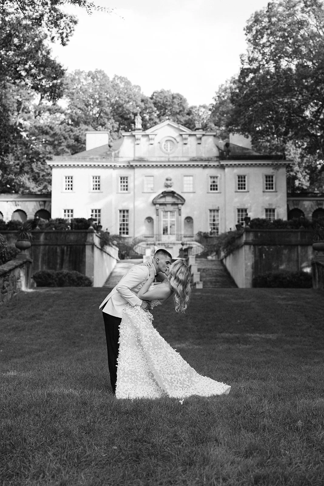A couple in wedding attire sharing a romantic moment on a lawn in front of a large, elegant mansion surrounded by trees.