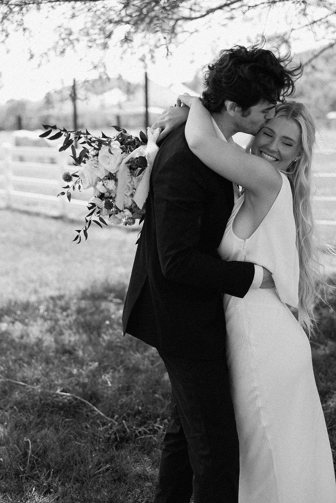 Black and white photo of a joyful bride and groom embracing outdoors, with the groom kissing the bride's cheek, holding a bouquet of flowers.