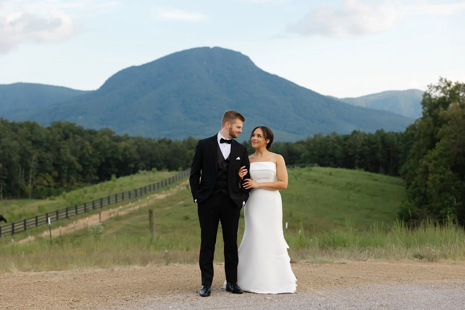 A couple in wedding attire standing on a dirt road with a mountain and green landscape in the background.