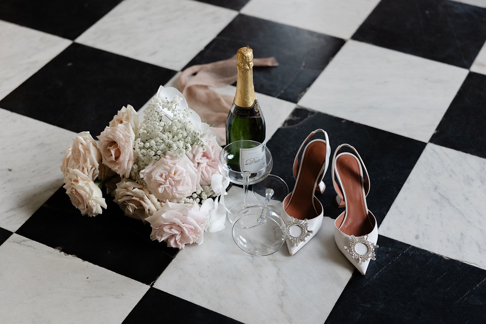 Wedding items on a black and white checkered floor, including a bouquet of pale pink and white flowers, a bottle of champagne, two empty wine glasses, and a pair of white bridal shoes with jewel embellishments.