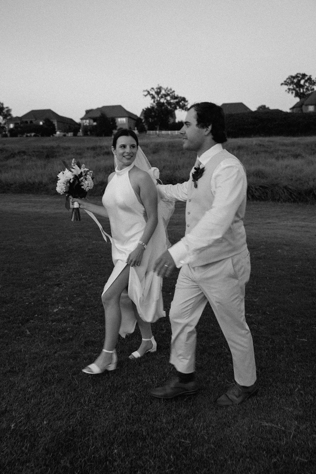 Black and white photo of a bride and groom walking outside on grass at sunset, with houses and trees in the background. The bride holds a bouquet and looks at the groom, who is smiling and looking at her.