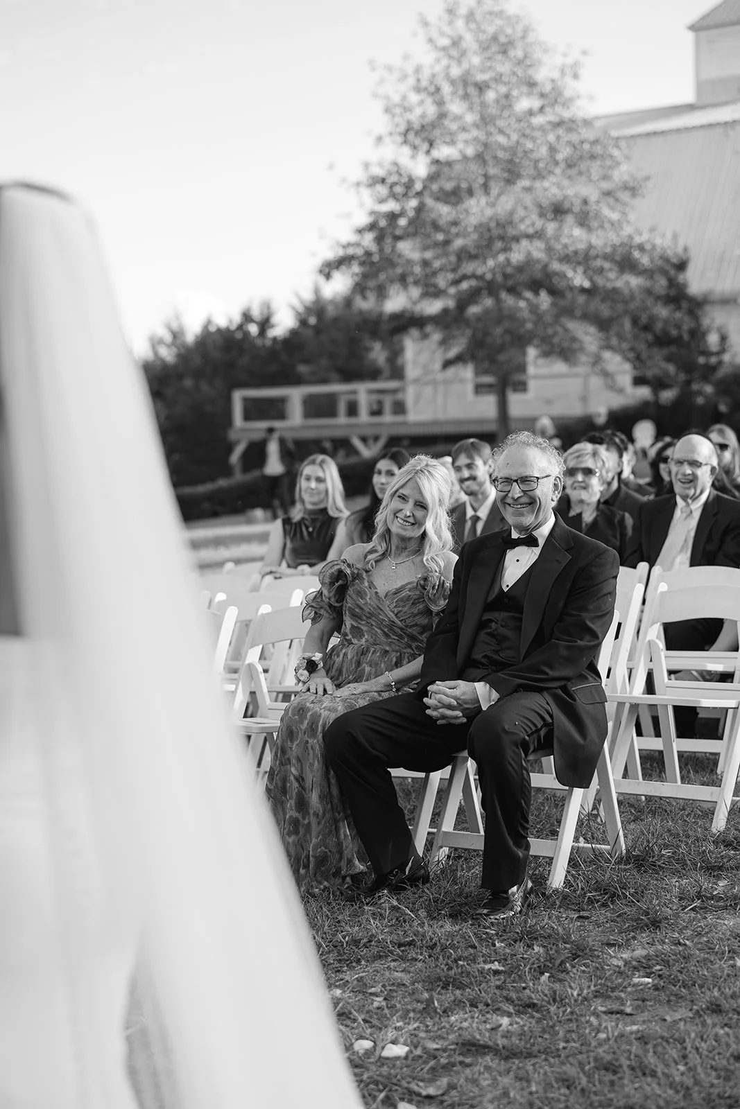 Black and white photo of wedding guests sitting outdoors, smiling and enjoying the ceremony, with a tree and house in the background.