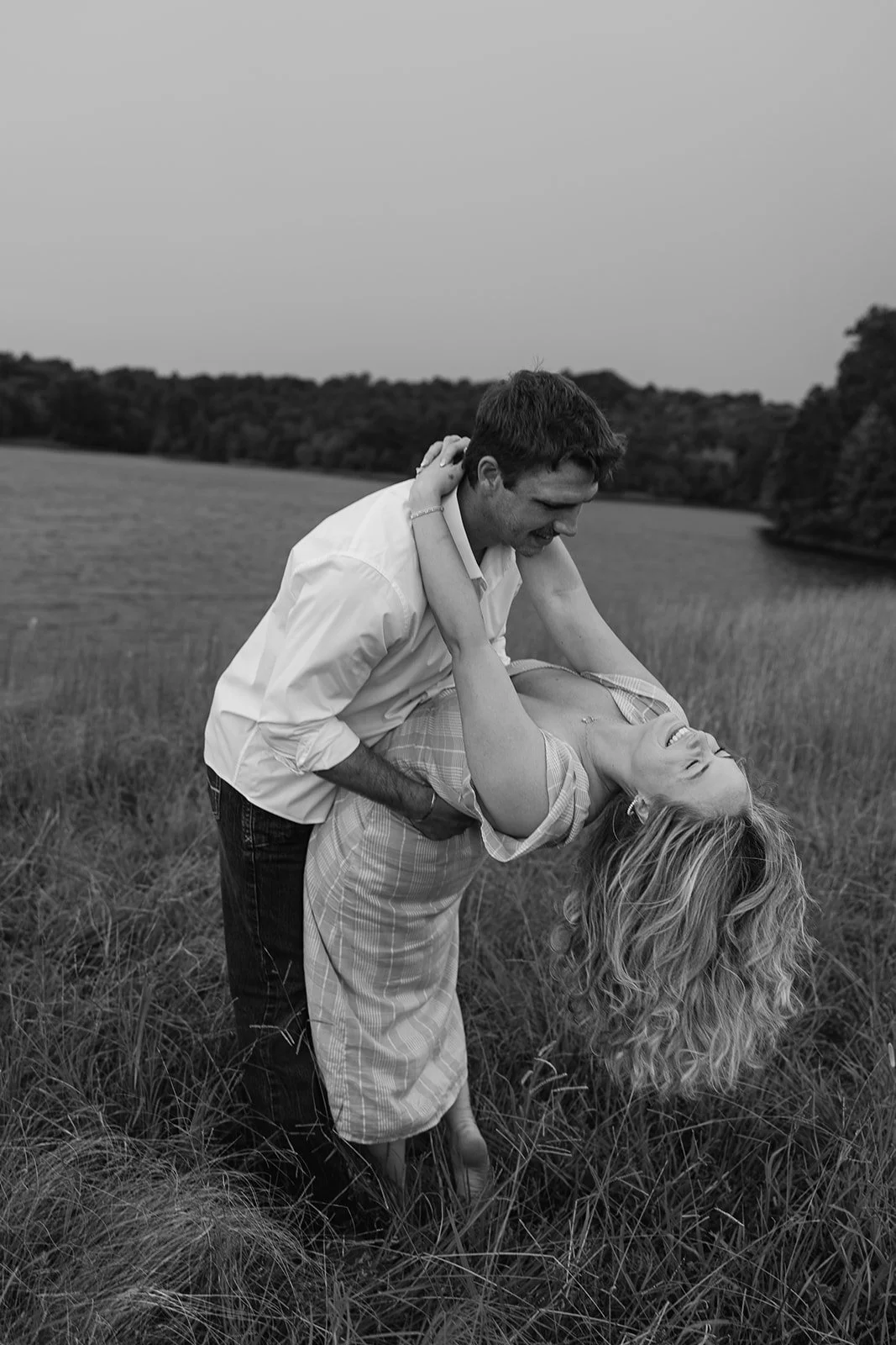 A black and white photo of a man and a woman dancing together outdoors near a lake, with the woman dipping backwards and smiling, and the man holding her, smiling as well.