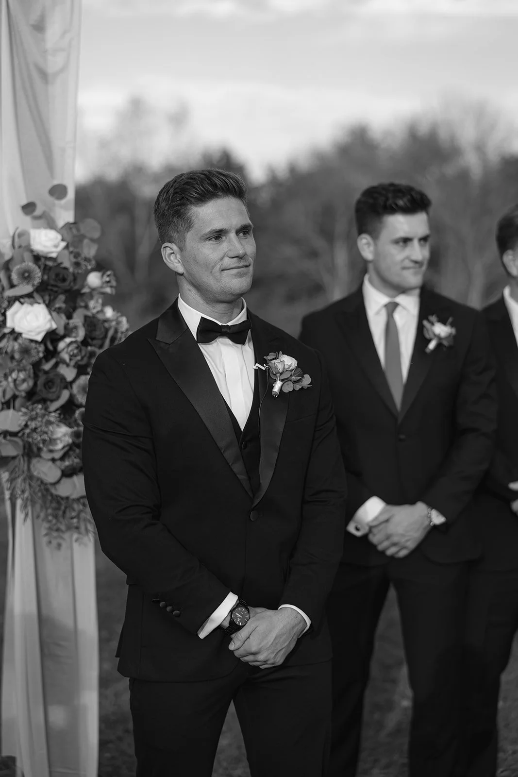 A black and white photo of a groom in a tuxedo with a bow tie and a boutonniere, standing outdoors during a wedding ceremony, with other men in suits visible in the background.