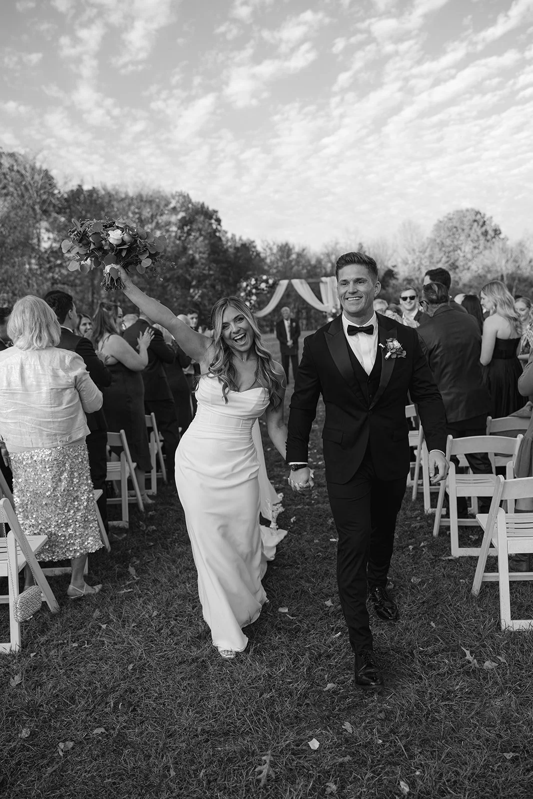 A bride and groom walking hand in hand at their wedding ceremony outdoors, smiling, with guests in the background.