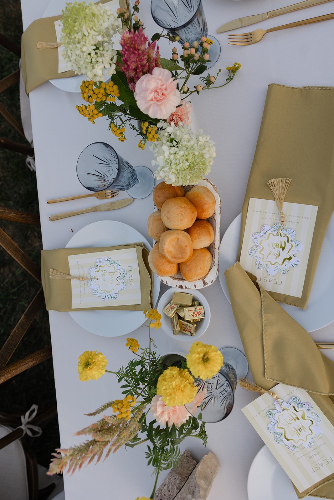 A table set for a meal with a white tablecloth, yellow napkins, and decorative plates. There are floral arrangements with pink, yellow, and white flowers, a bouquet of bread rolls in a rectangular dish, small wrapped candies, and a glass of water wit