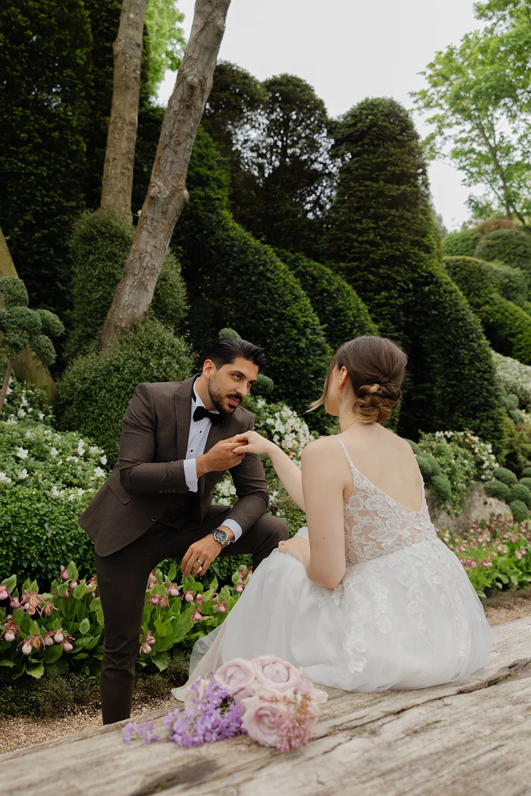 A groom kneeling and holding the hand of a bride who is sitting outdoors in a garden with greenery and flowers.