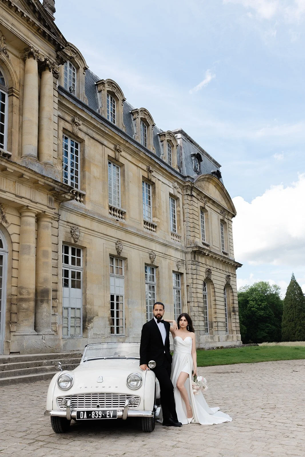 A bride and groom in wedding attire standing next to a vintage white car outside a grand historic building with large windows and ornate stone detailing, under a partly cloudy sky.