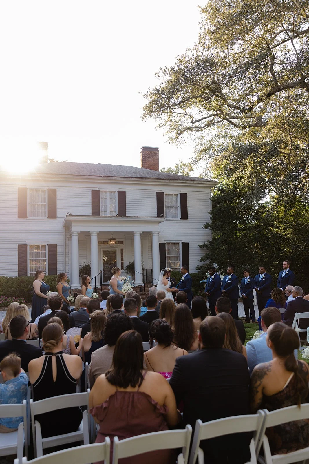 A wedding ceremony outside a large white house with black shutters, with the bride and groom exchanging vows underneath a canopy, surrounded by bridesmaids and groomsmen, with guests seated in front.