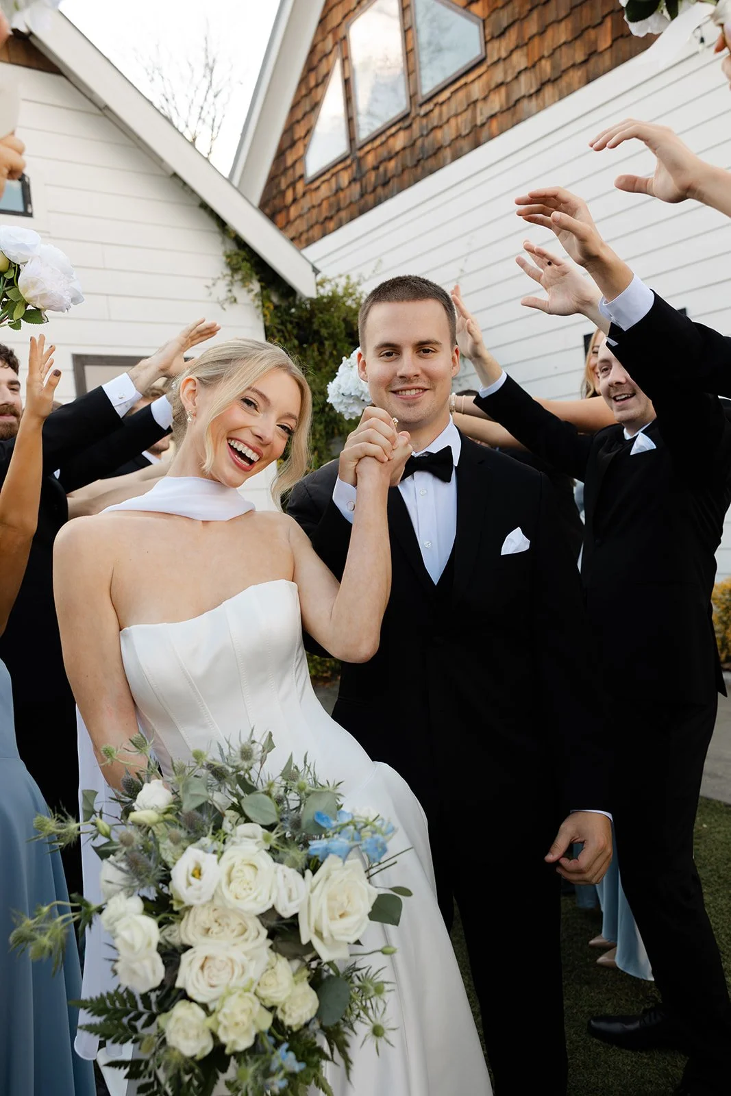 A bride and groom are celebrating their wedding outdoors with friends. The bride is wearing a white wedding dress and holding a bouquet of white and blue flowers. The groom is in a black tuxedo. People around them are raising their hands in celebrati
