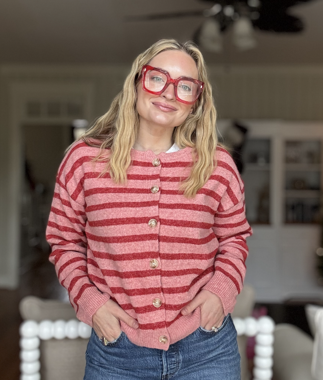 Woman with long blonde hair wearing large red glasses, a pink and red striped cardigan with pearl buttons, and jeans, standing indoors with hands in pockets.