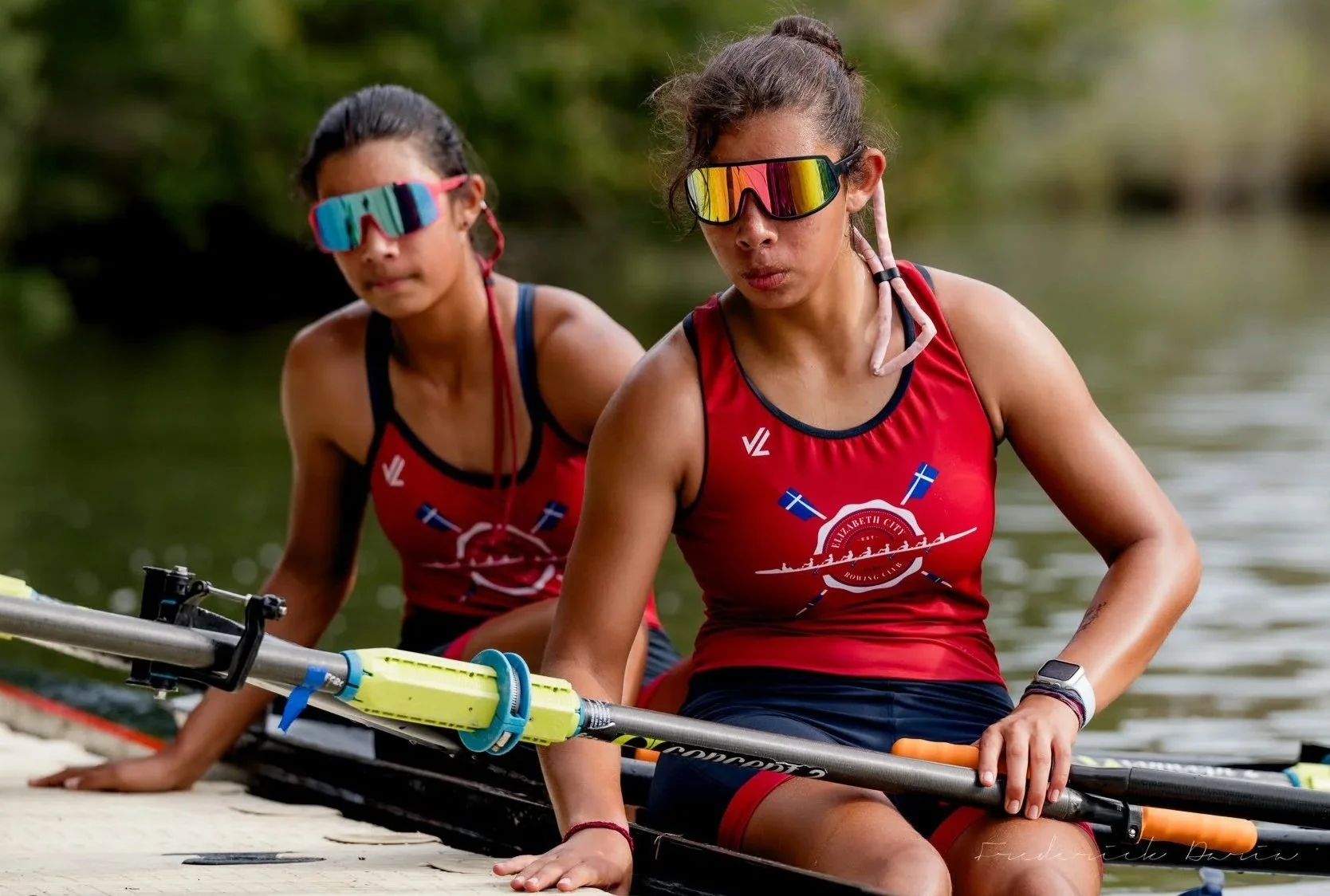 Two young women in Elizabeth City Rowing Club team athletic wear, sitting on a boat with paddles, near a body of water, wearing sunglasses and red tank tops.